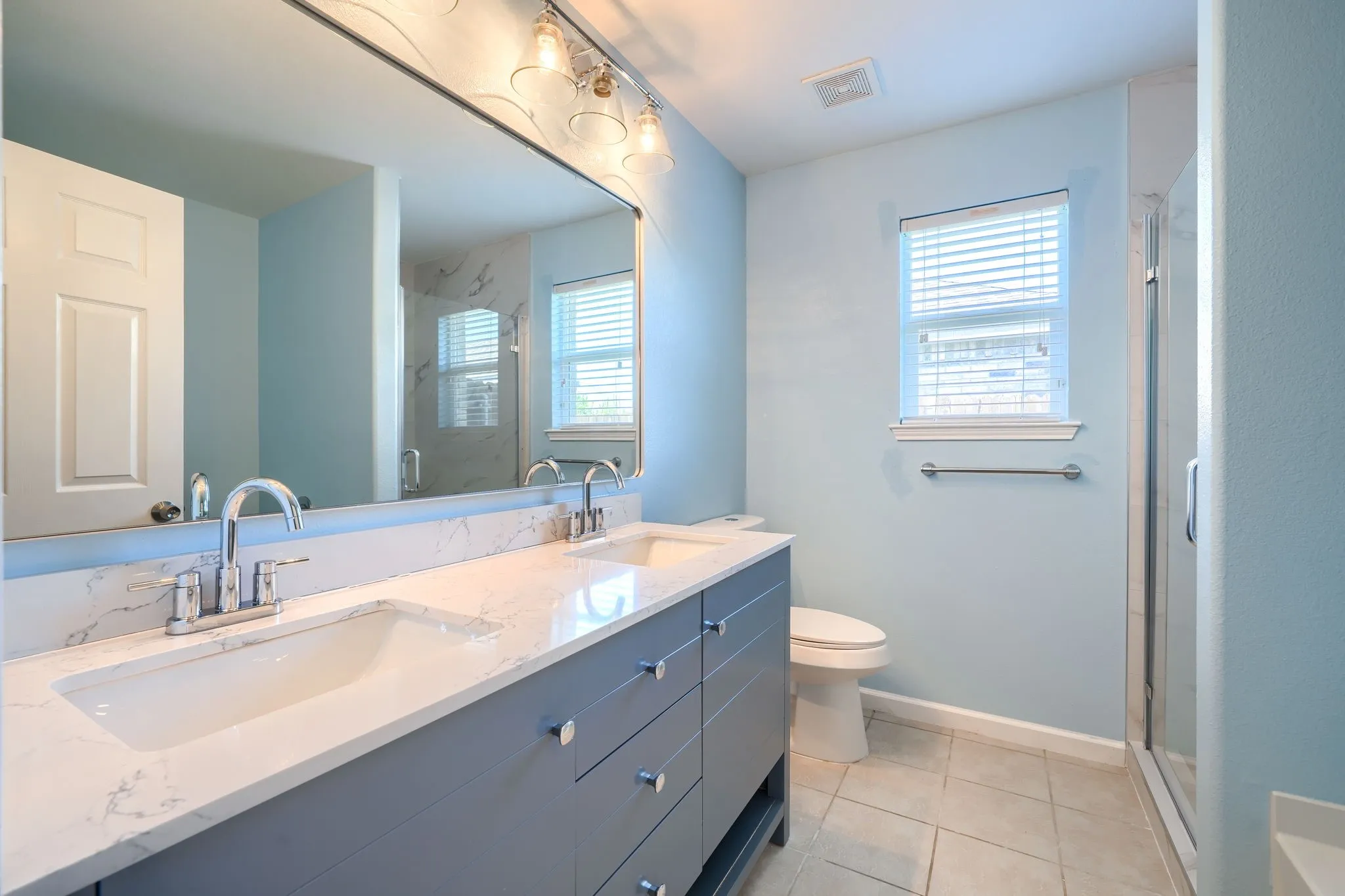 Bathroom vanity featuring dual under-mount sinks with chrome fixtures, a broad mirror with overhead lighting, and a white countertop