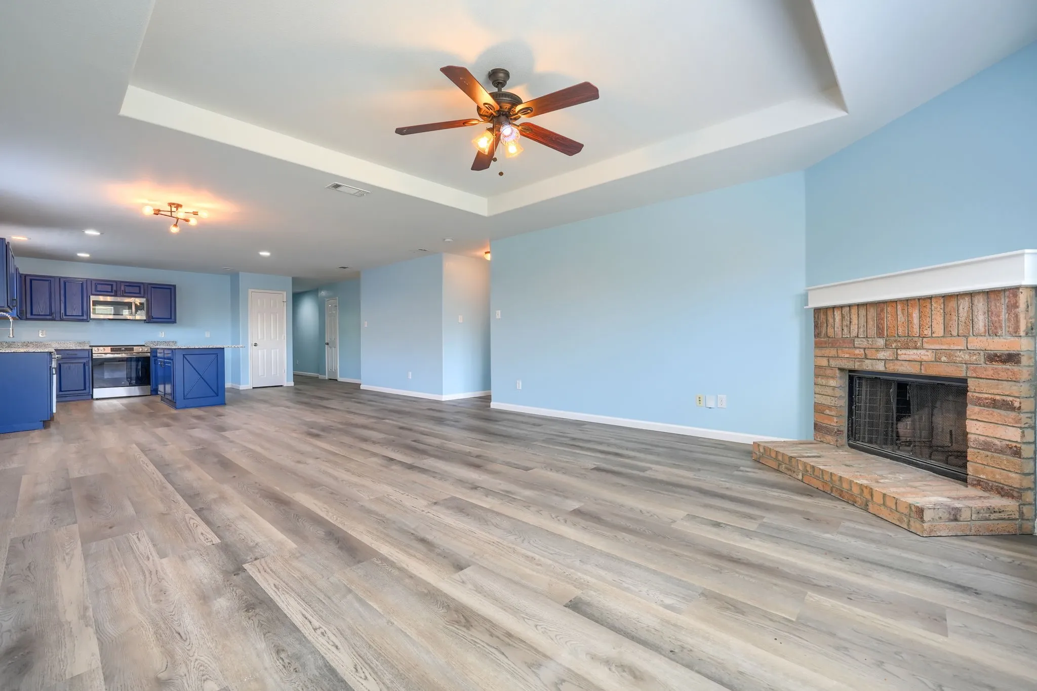 Spacious open concept room featuring wood-finish flooring, a tray ceiling with a ceiling fan, and a brick fireplace