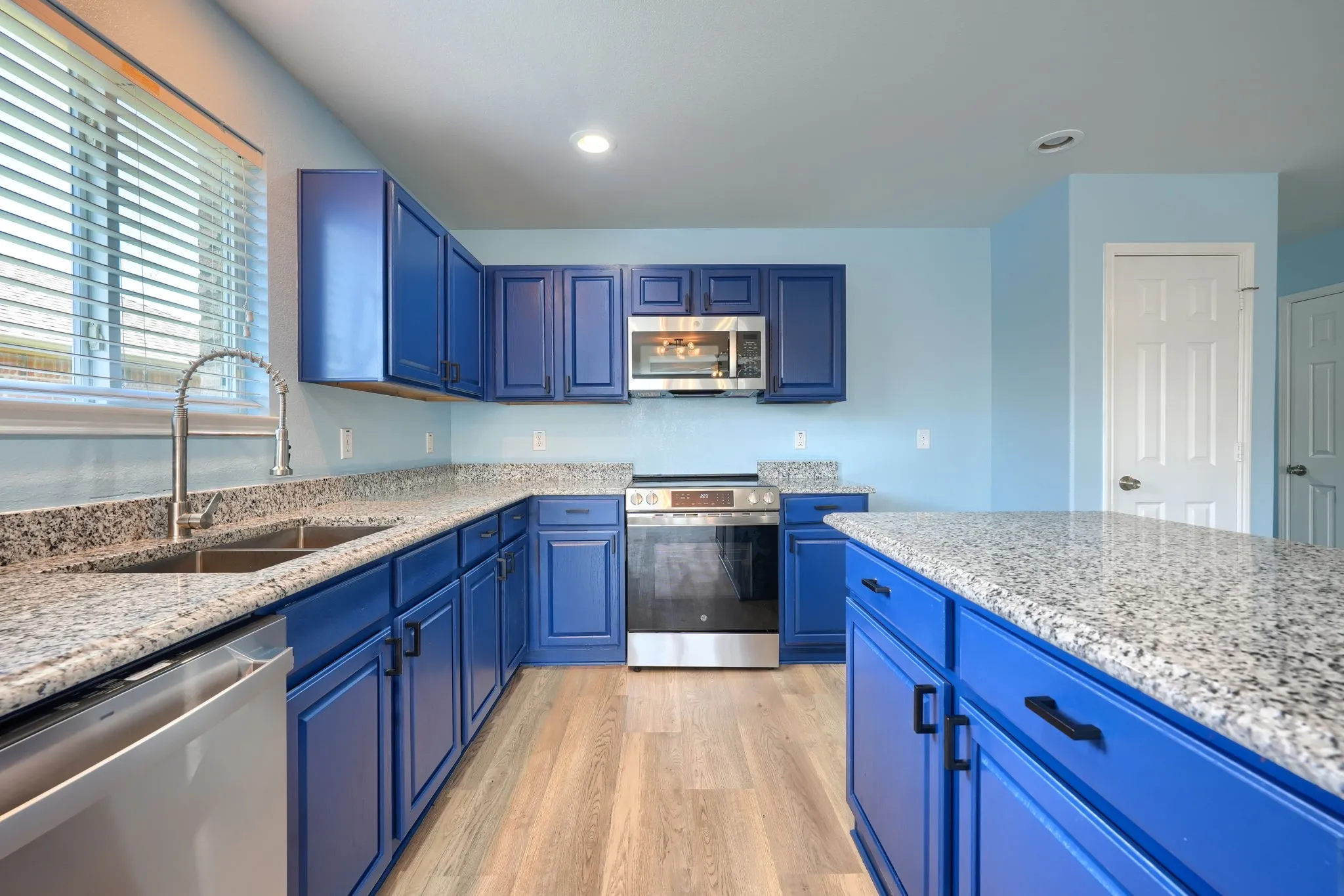 Bright kitchen featuring blue cabinetry, light wood-finish flooring, stainless steel appliances, a double basin sink, and a center island with a granite-style countertop