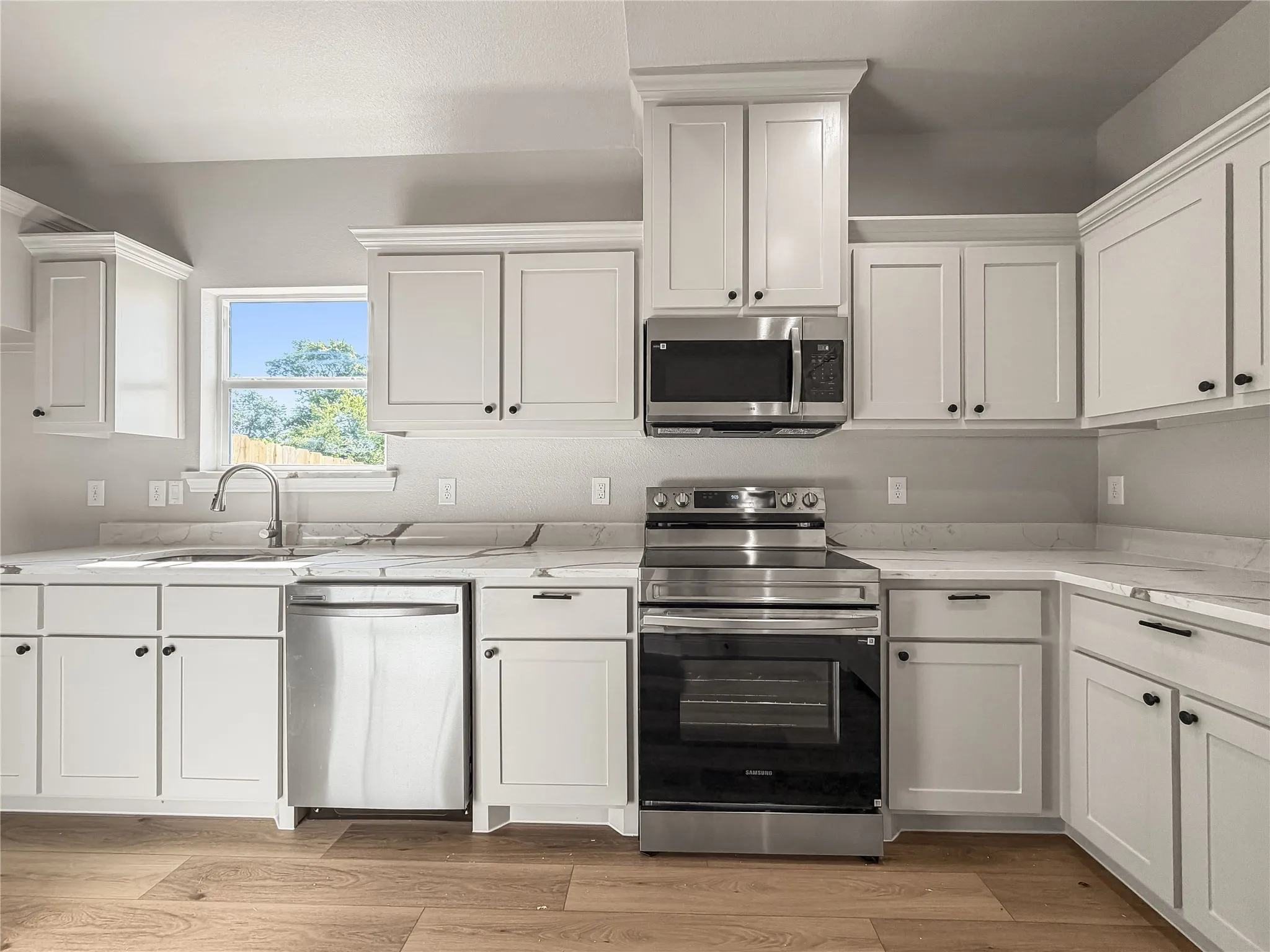 Bright kitchen featuring white shaker-style cabinetry, stainless steel appliances, light-toned countertops, a stainless steel sink with a gooseneck faucet, and wood-finish flooring