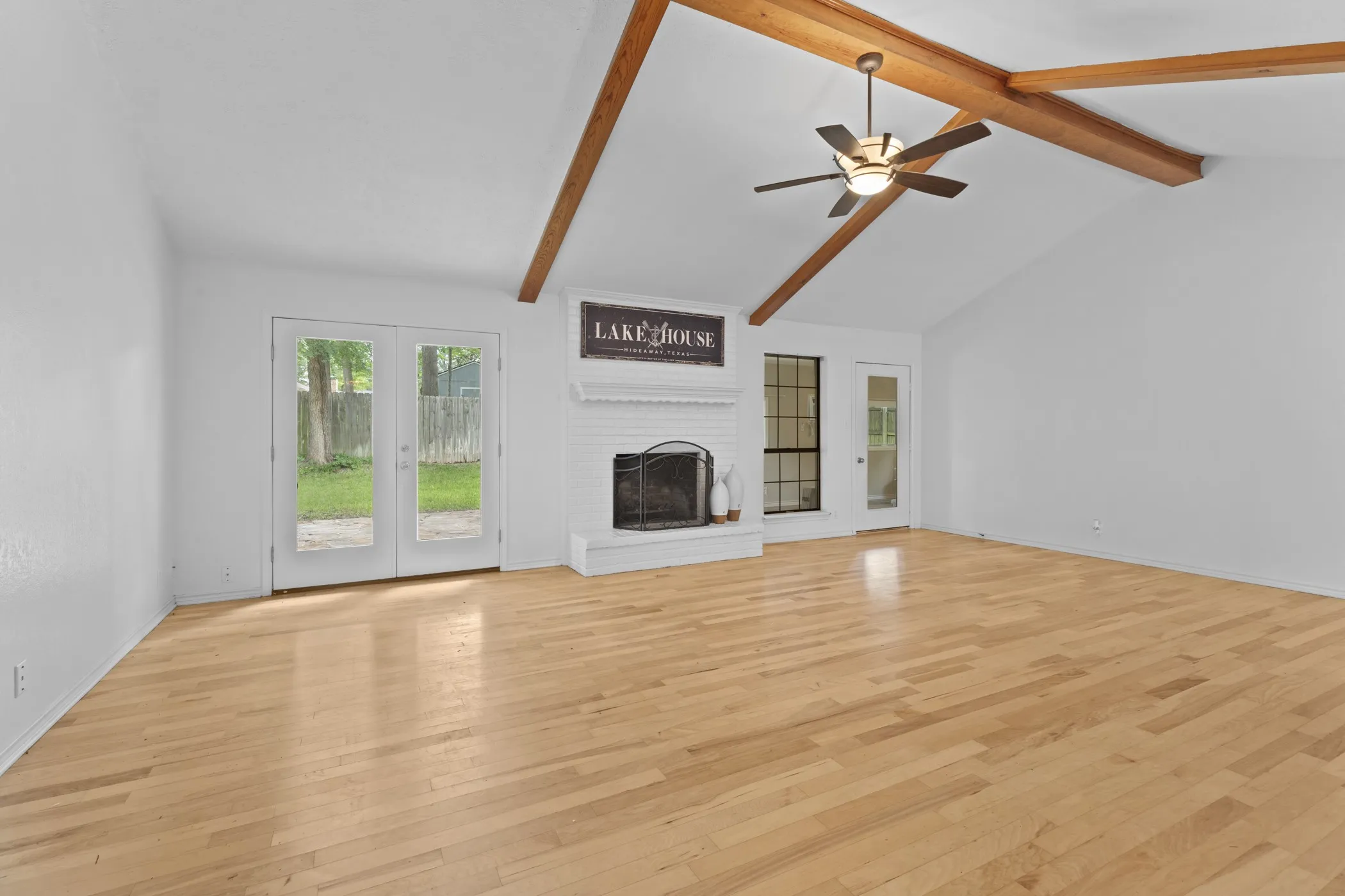 Vaulted ceiling with exposed wood beams, wood-finish flooring, white brick fireplace with mantel, ceiling fan, and double glass-paneled French doors opening to an exterior area