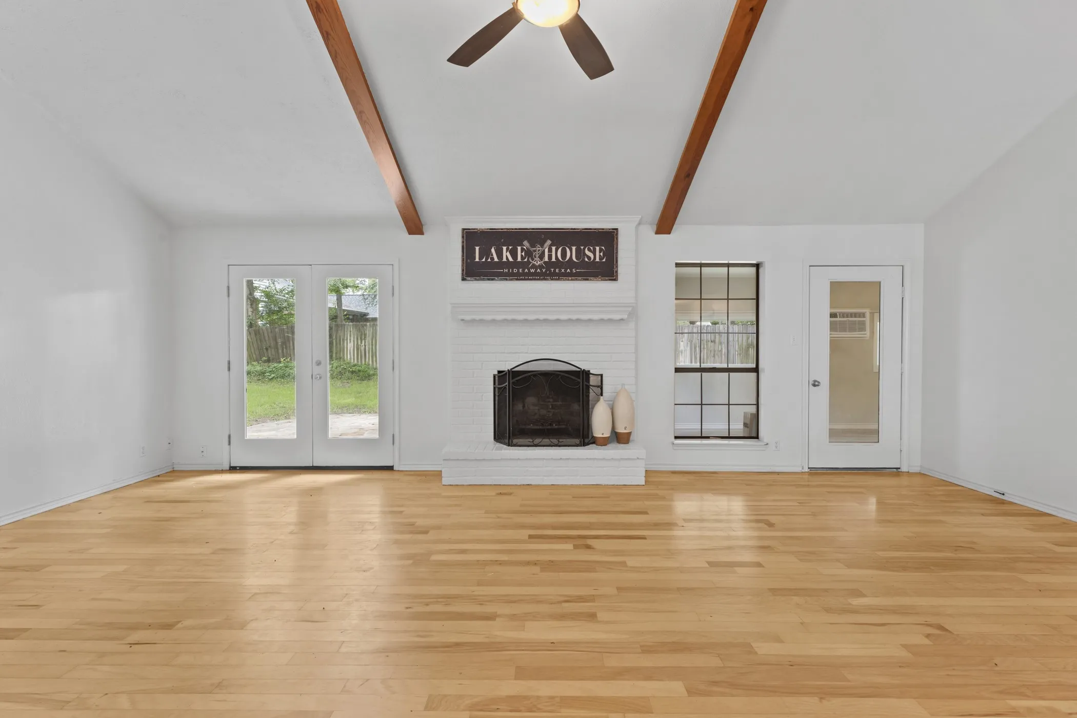 Spacious living area featuring light wood-finish flooring, a white brick fireplace with a black fire screen, vaulted ceilings with exposed wood beams, and a ceiling fan