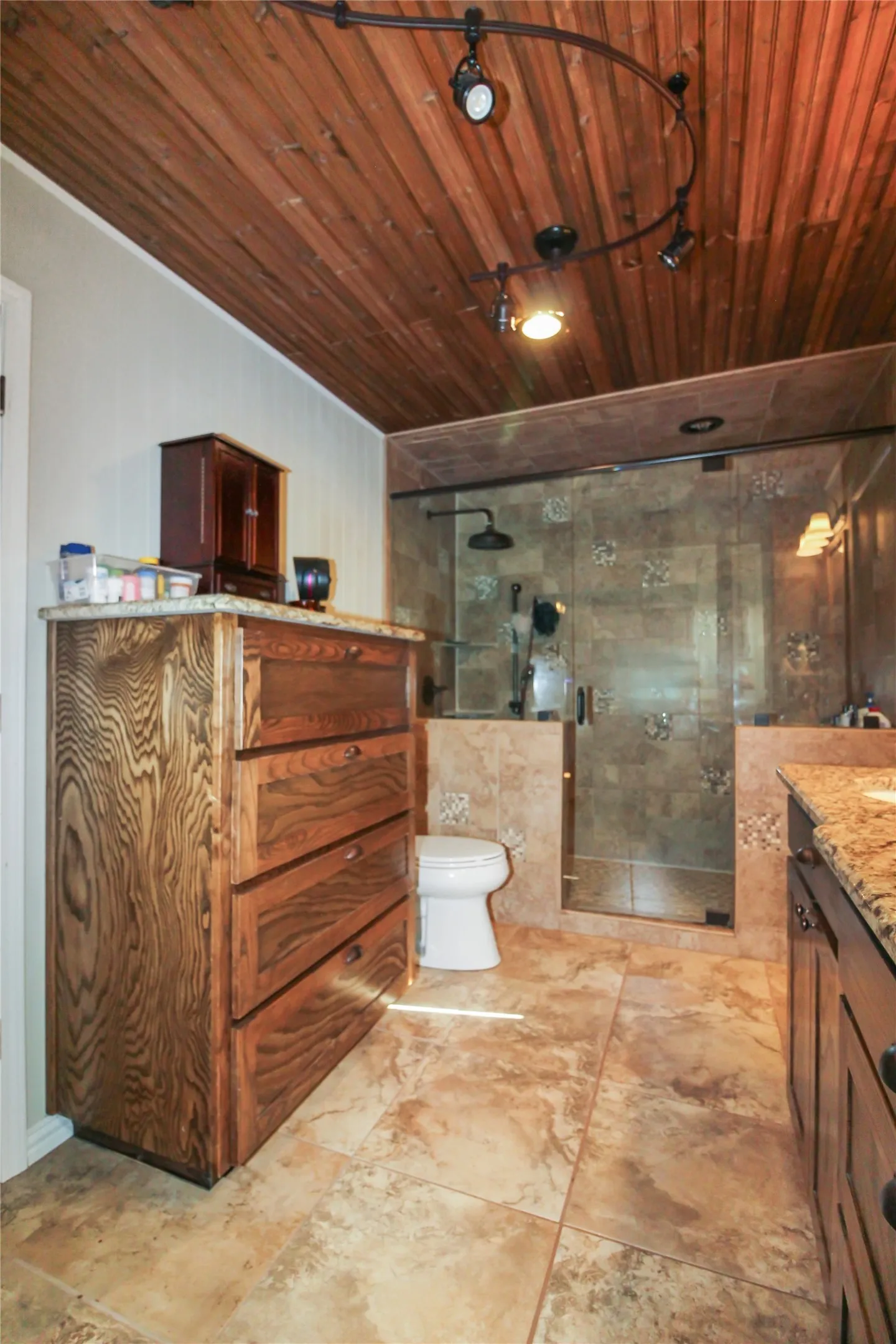 Bathroom featuring a wood-panel ceiling with track lighting, an enclosed shower with tile surround, a built-in wood vanity with a granite-finish countertop, and large format floor tiling