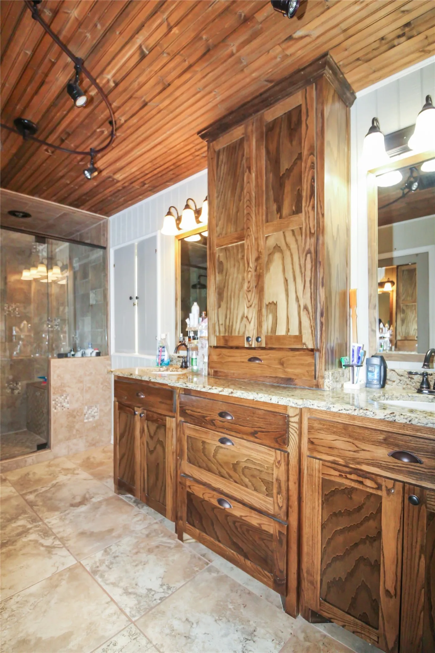 Spacious bathroom featuring a wood-planked ceiling with track lighting, dual wood-finish vanity with granite-style countertops, wood-finish cabinetry, and a walk-in shower with tiled walls and a glass enclosure