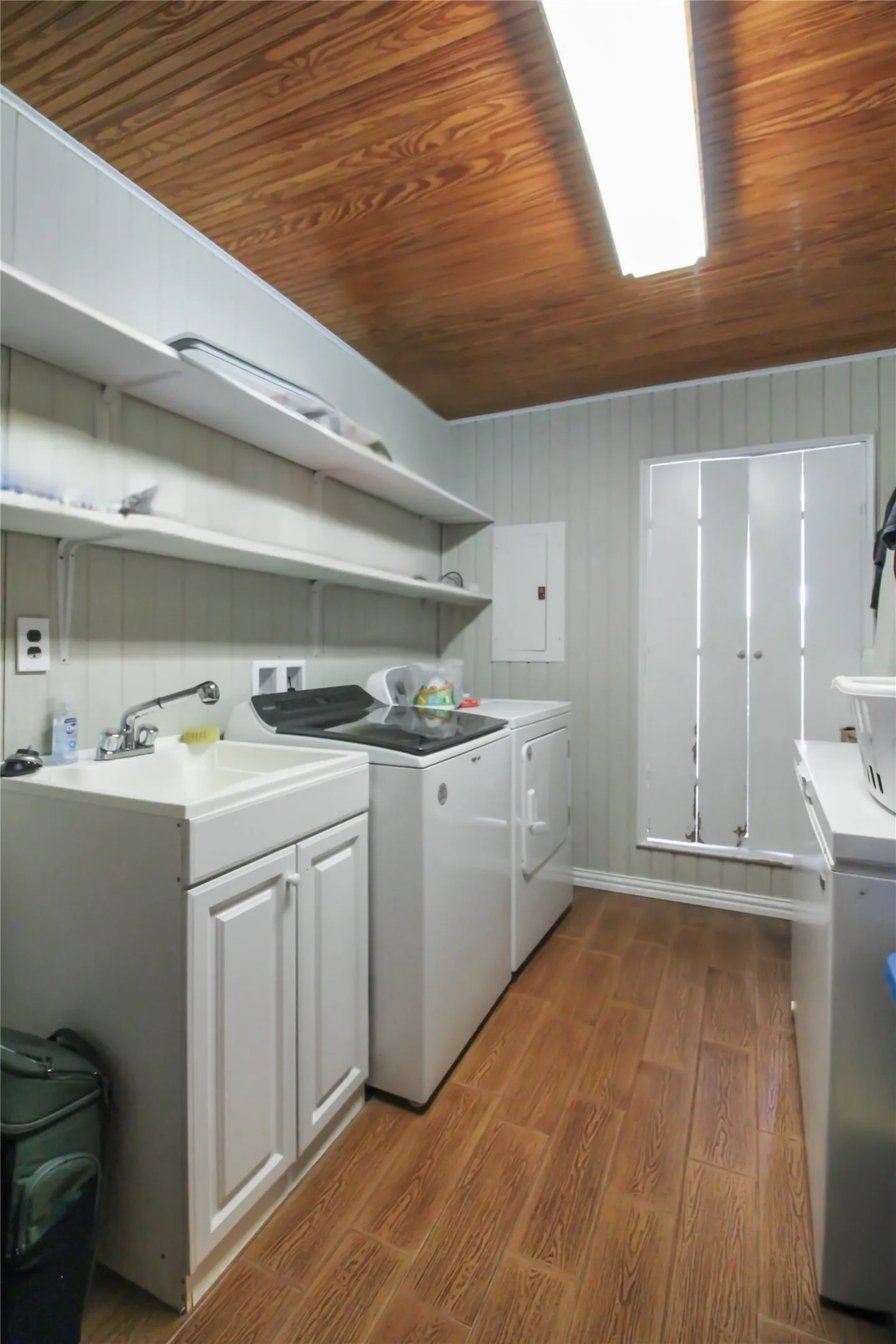 Laundry area featuring wood-finish flooring, beadboard wall paneling, a utility sink with white cabinetry, and built-in shelving
