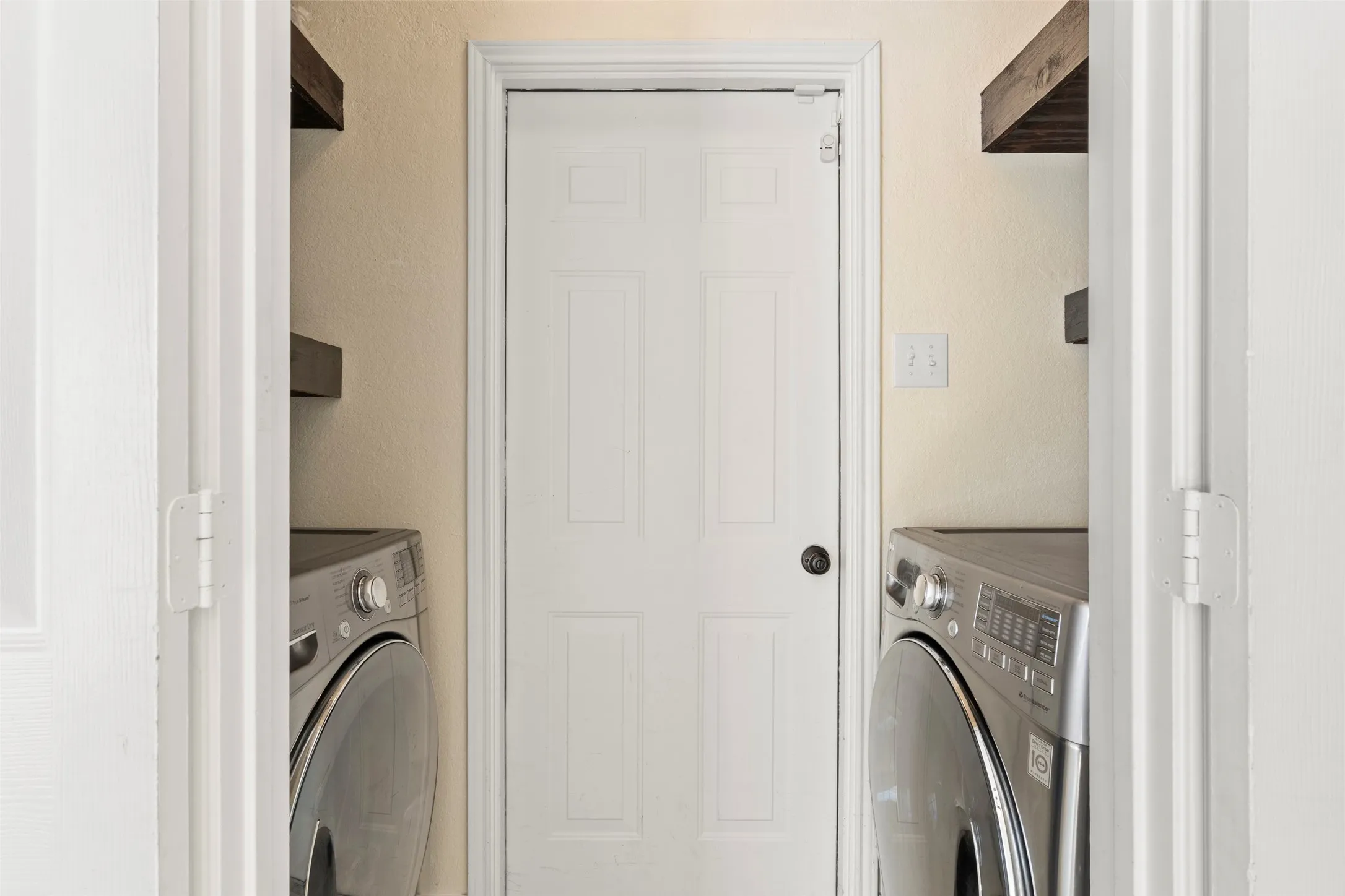 Dedicated laundry area featuring a central white paneled door, light-toned walls, and wood-finish floating shelves for storage