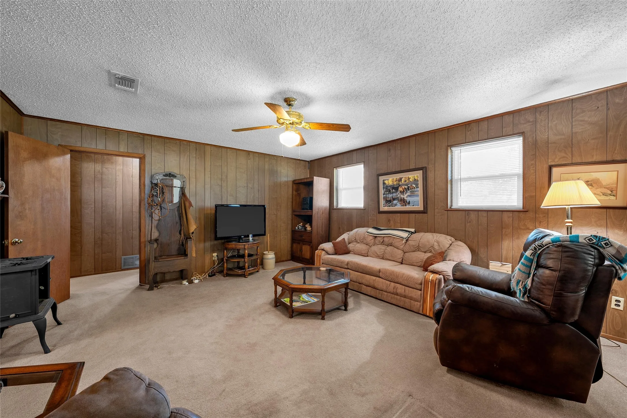 Carpeted living space featuring wood paneling, multiple windows with blinds, a ceiling fan with integrated lighting, and a dark-toned wood stove