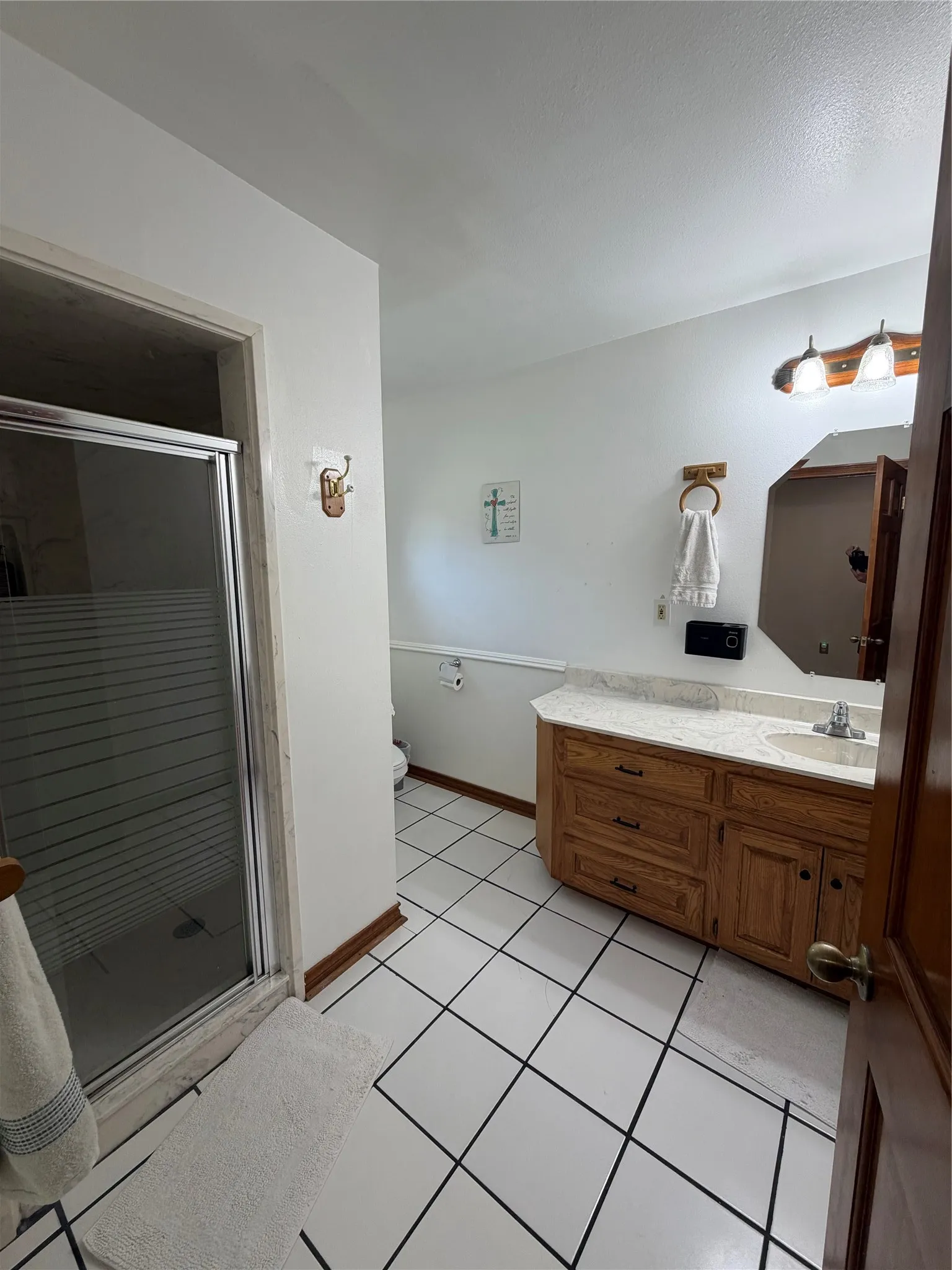 Bathroom featuring a glass-enclosed shower, white tile flooring with dark grout, a wood-finish vanity with ample storage, a countertop, and a built-in sink with a chrome faucet
