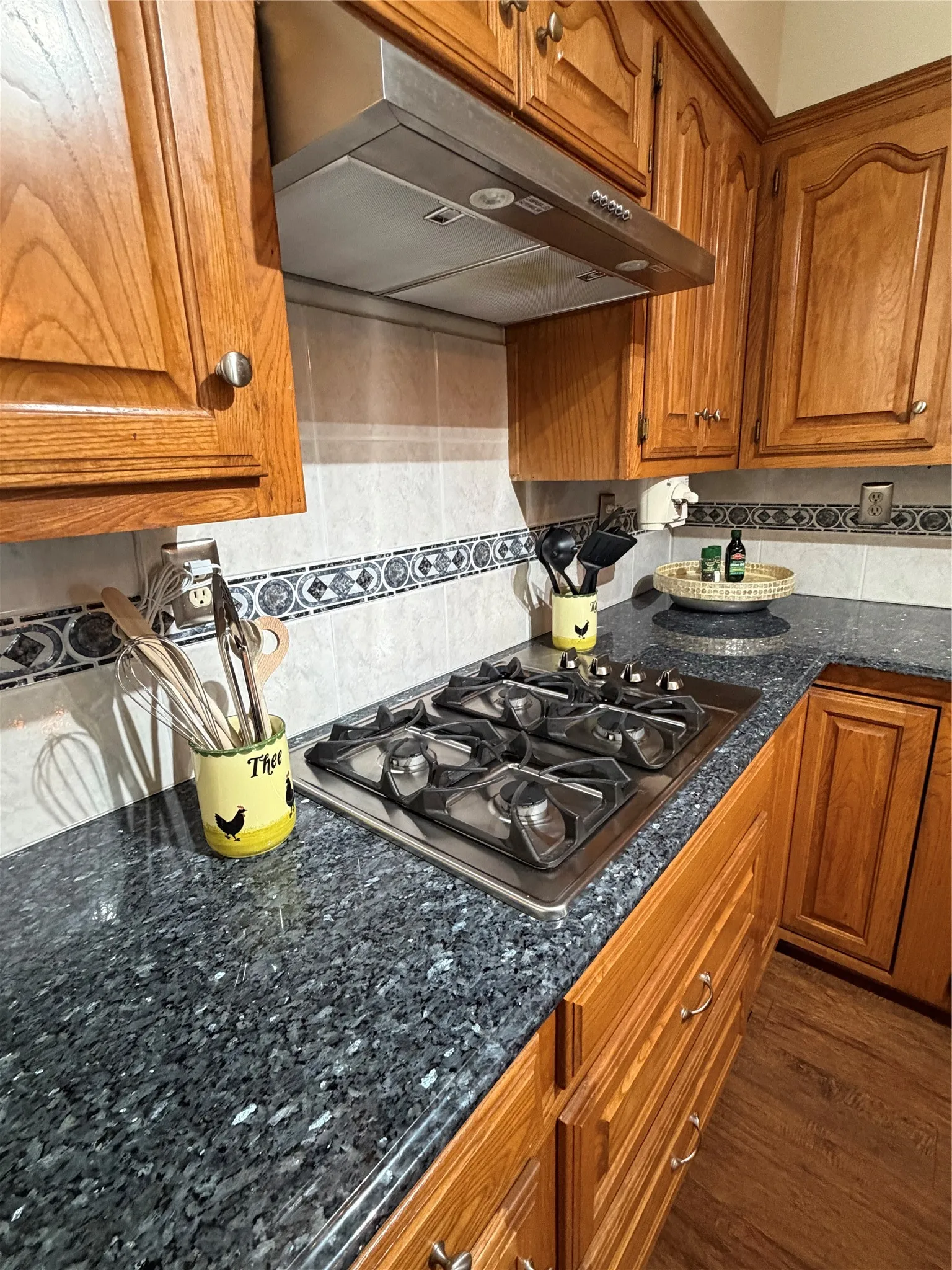 Kitchen featuring a built-in stainless steel gas cooktop, a coordinating stainless steel range hood, and dark granite countertops