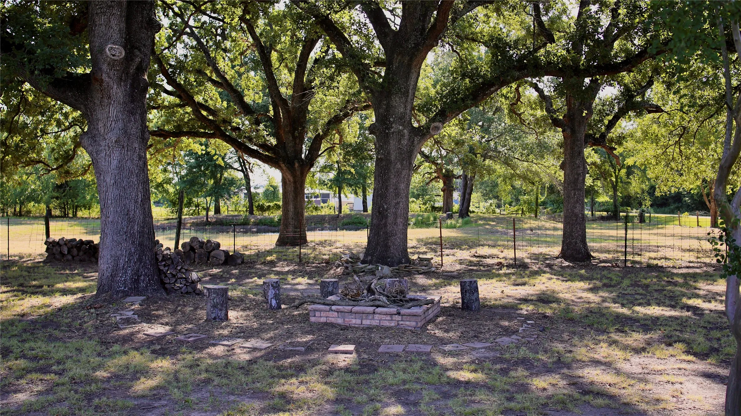 Spacious outdoor area featuring mature trees providing ample shade, a brick-lined fire pit, and a partial perimeter fence
