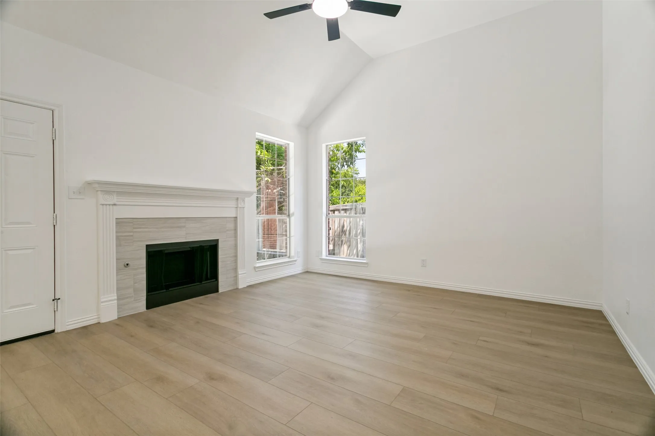 Bright living space featuring a vaulted ceiling with ceiling fan, wood-finish flooring, and a fireplace with a white mantel and light-toned tile surround
