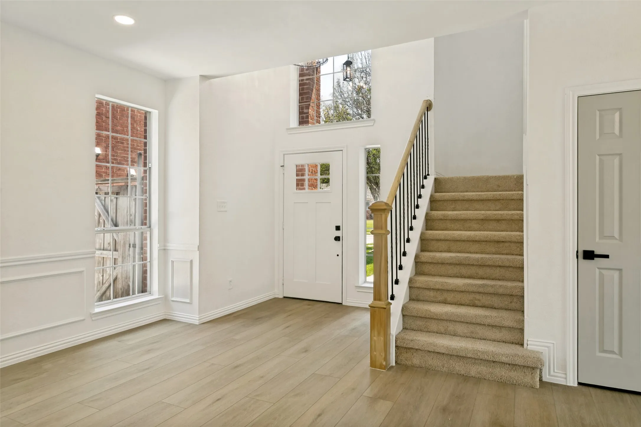 Two-story foyer featuring wood-finish flooring, a carpeted staircase with wood and iron banister, a white paneled entry door with frosted glass inserts, and multiple windows including a tall fixed window with decorative trim