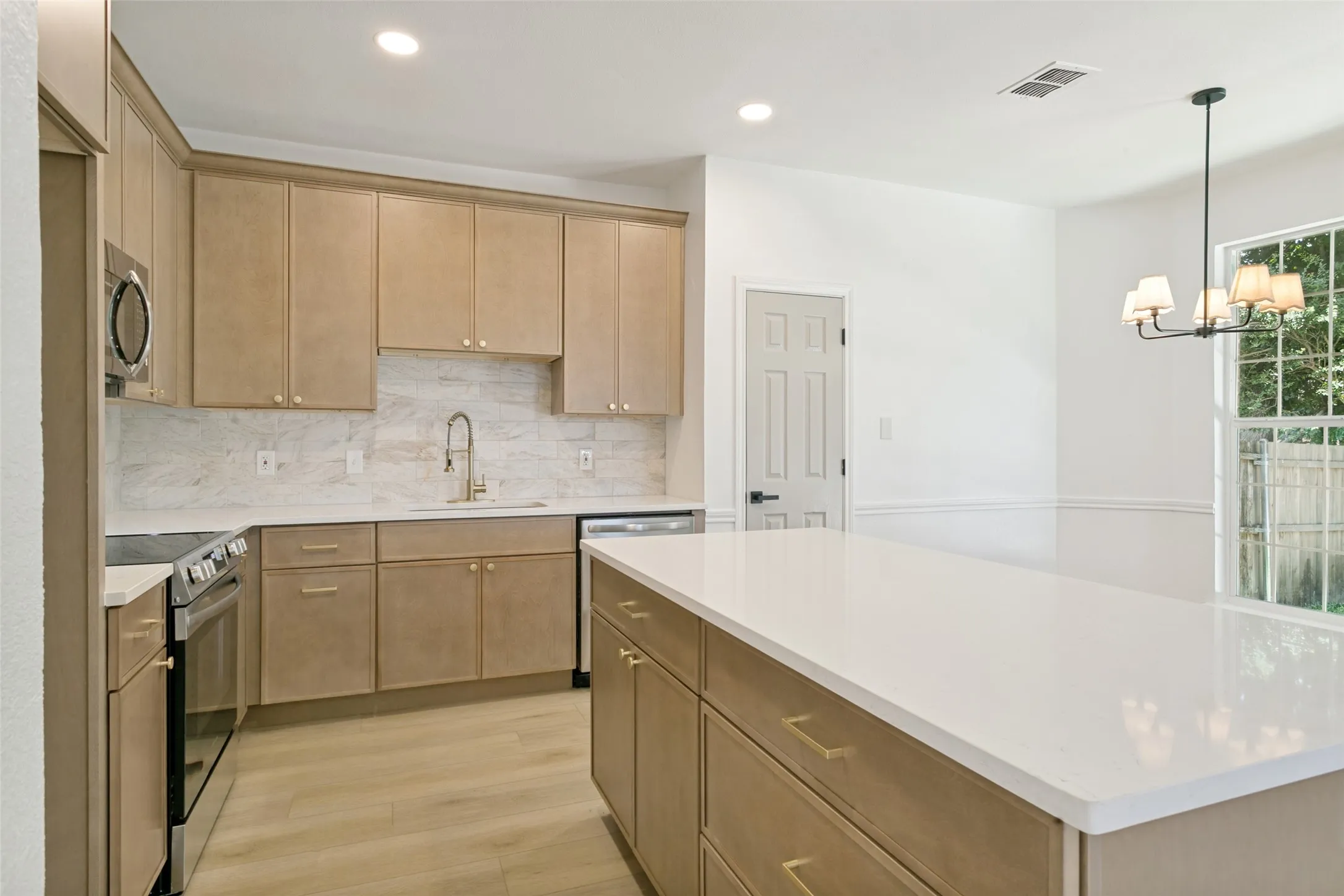 Modern kitchen featuring a large center island with a white countertop, light wood-finish cabinetry, stainless steel appliances, a white tile backsplash, and wood-finish flooring