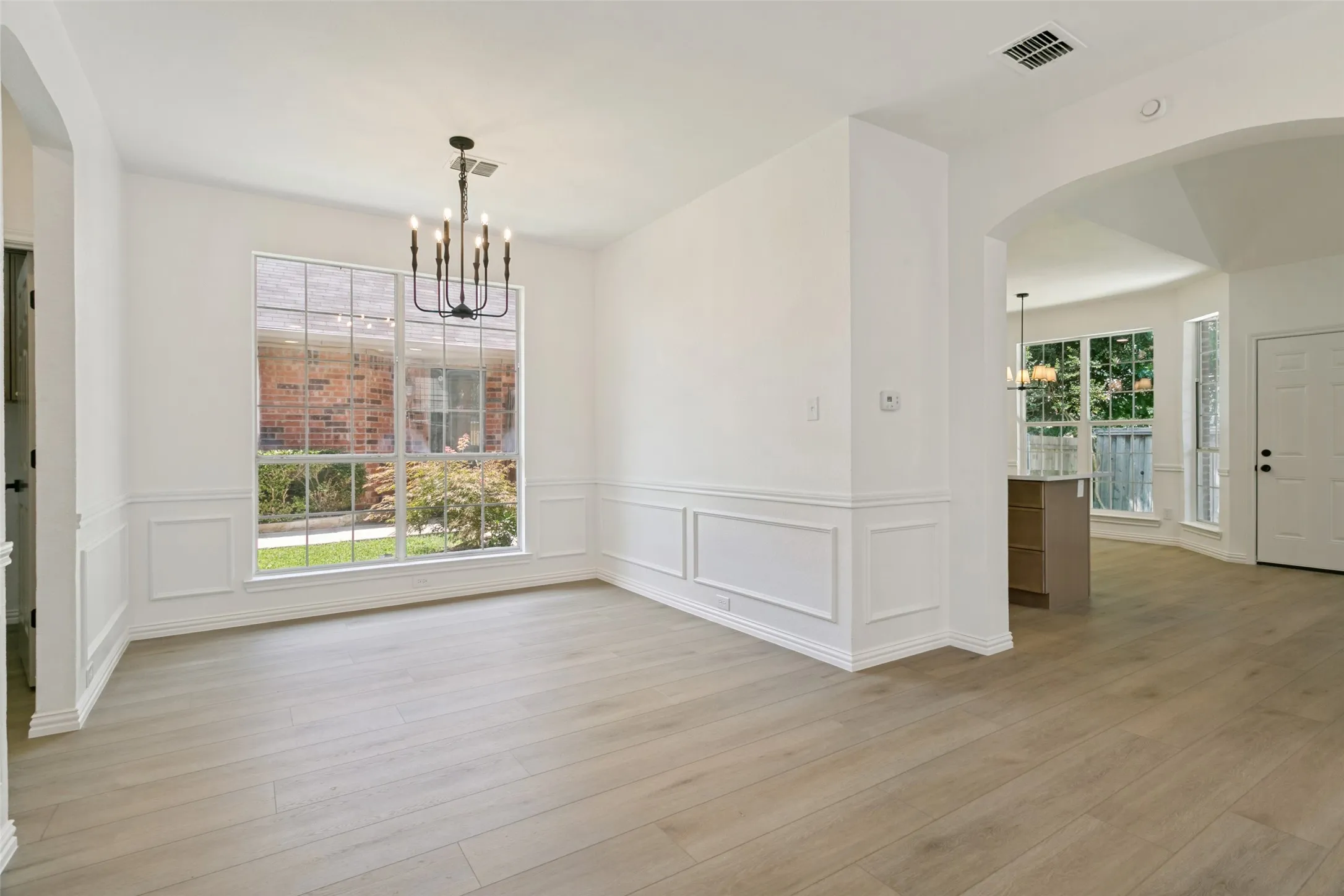 Light wood-finish flooring throughout, featuring a modern black chandelier, white wainscoting, and a large window overlooking exterior landscaping
