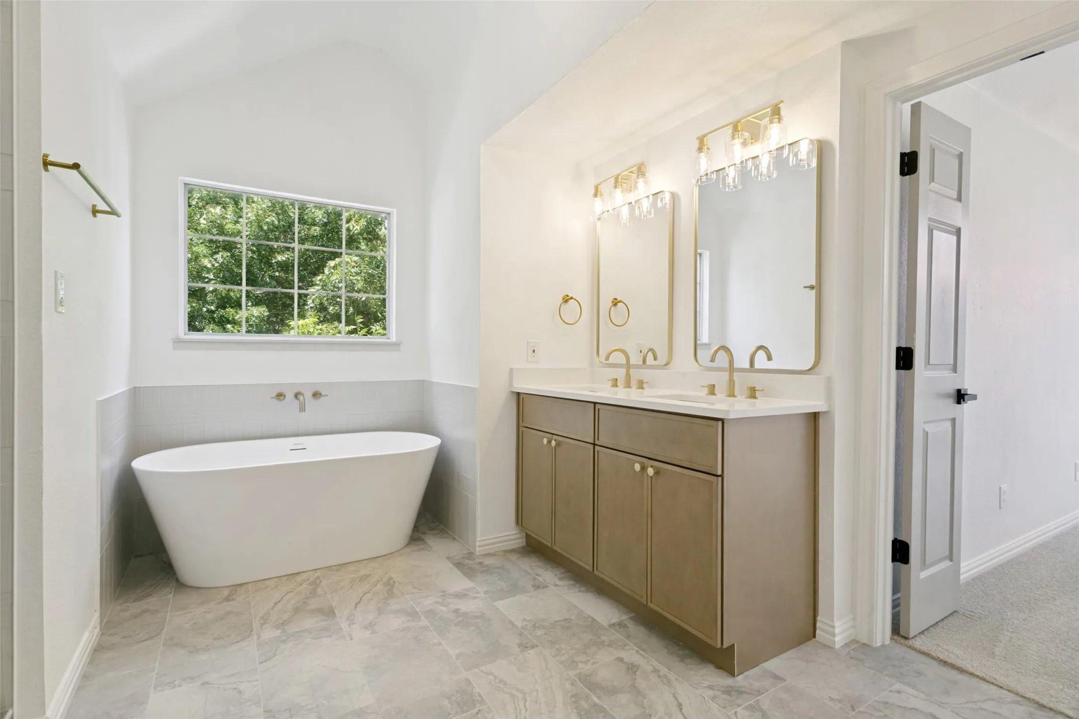 Freestanding soaking tub with wall-mounted faucet, dual vanity with undermount sinks, frameless mirrors with decorative lighting, vaulted ceiling, and large window
