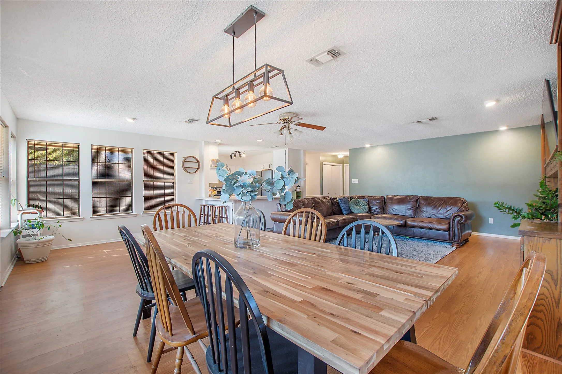 Expansive open-concept living area featuring a prominent wood-finish dining table, wood-finish flooring, a contemporary linear chandelier, and ample natural light from multiple windows