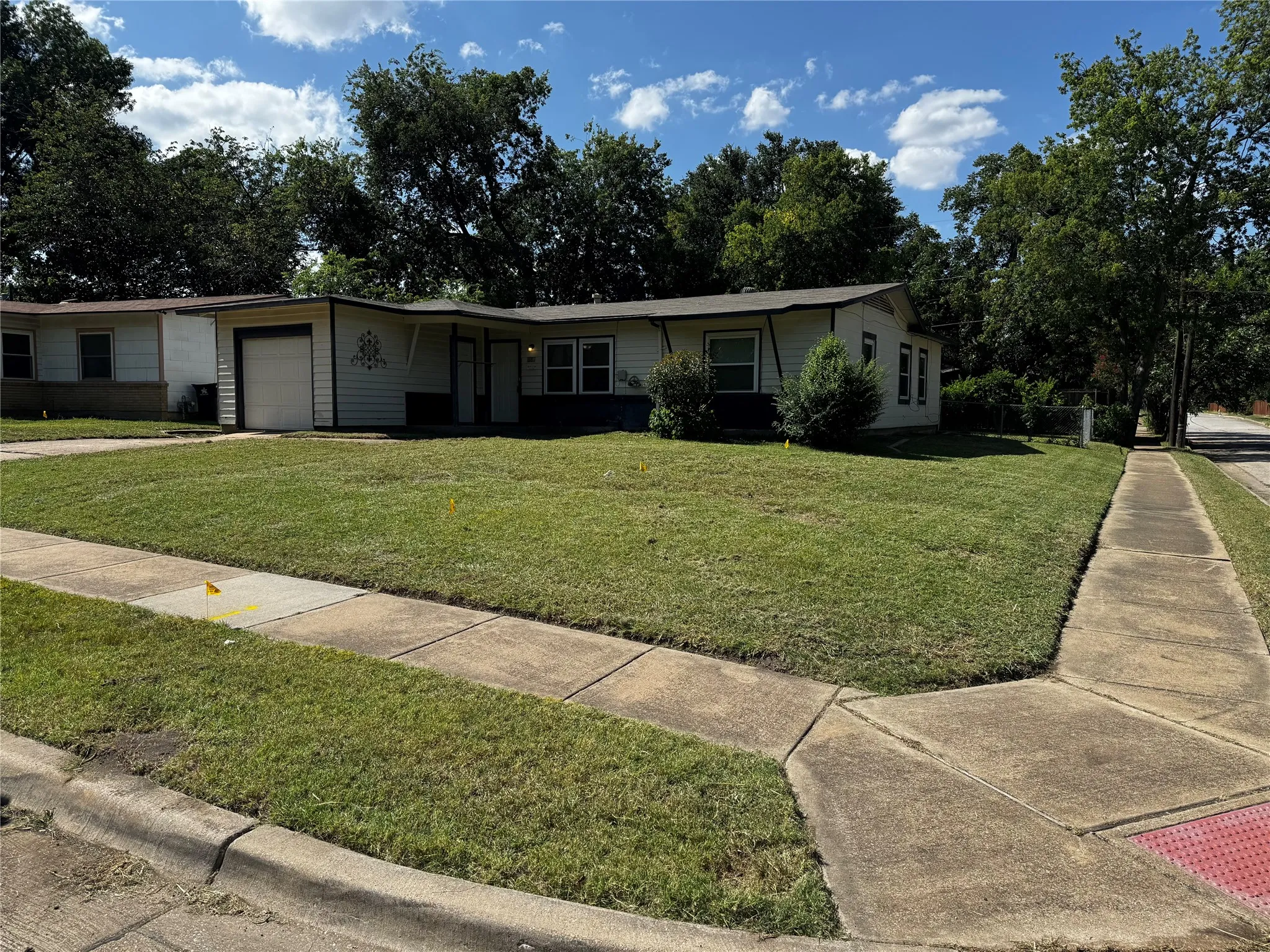 Corner lot property featuring a white exterior, a front-facing garage door, mature trees, and a concrete sidewalk
