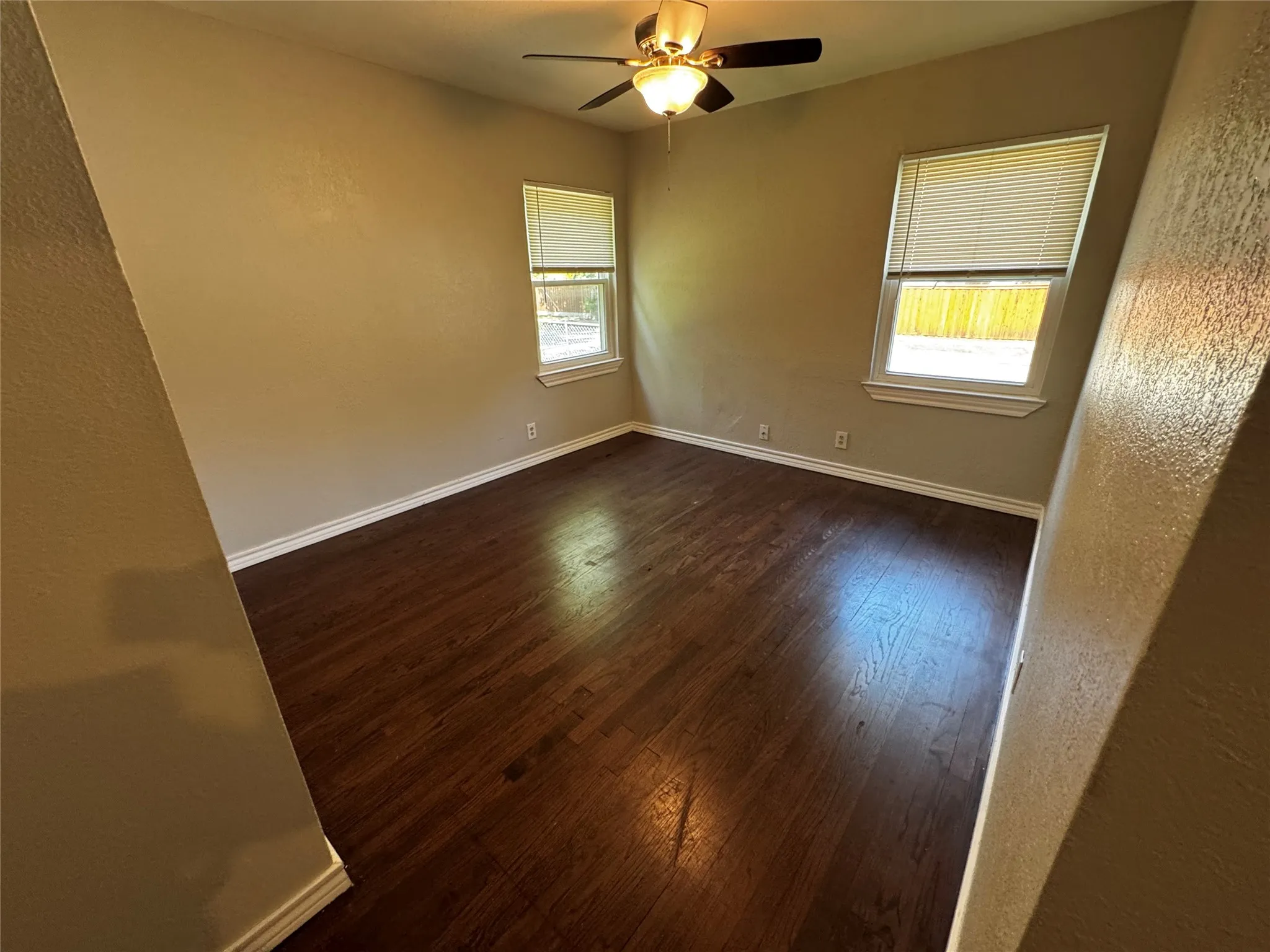 Spacious room featuring wood-finish flooring, neutral-toned walls, two windows with blinds, a ceiling fan with integrated lighting, and white baseboards