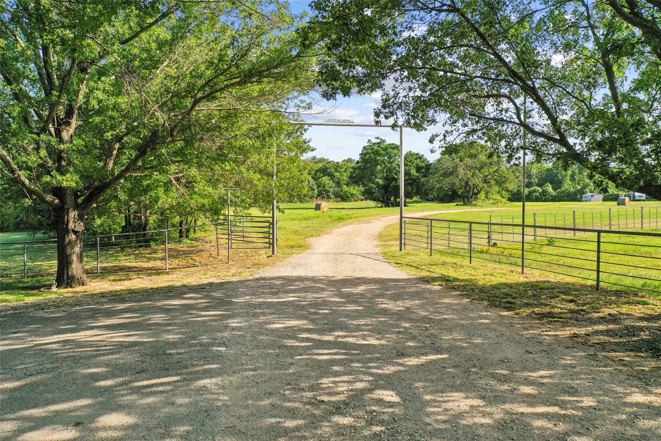Gravel driveway entry framed by mature trees, featuring a metal gate and fencing bordering expansive green pastures