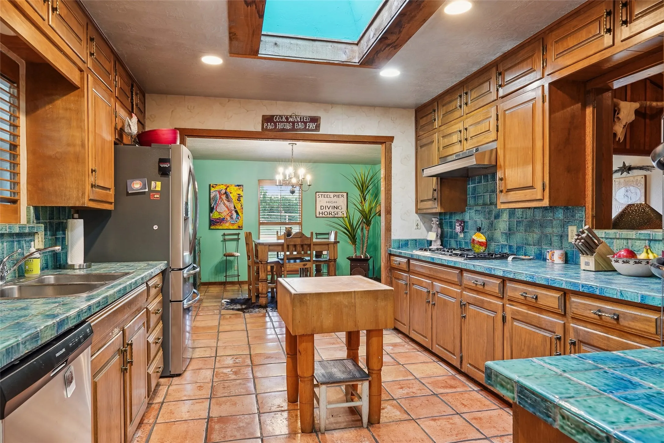 Open-concept kitchen featuring wood cabinetry, integrated appliances, ceramic tile flooring, a skylight, and vibrant tile countertops and backsplash