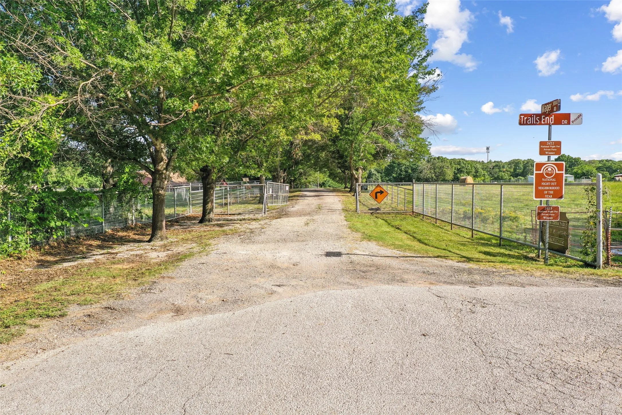 Gated entrance to a gravel drive, bordered by mature trees and chain-link fencing