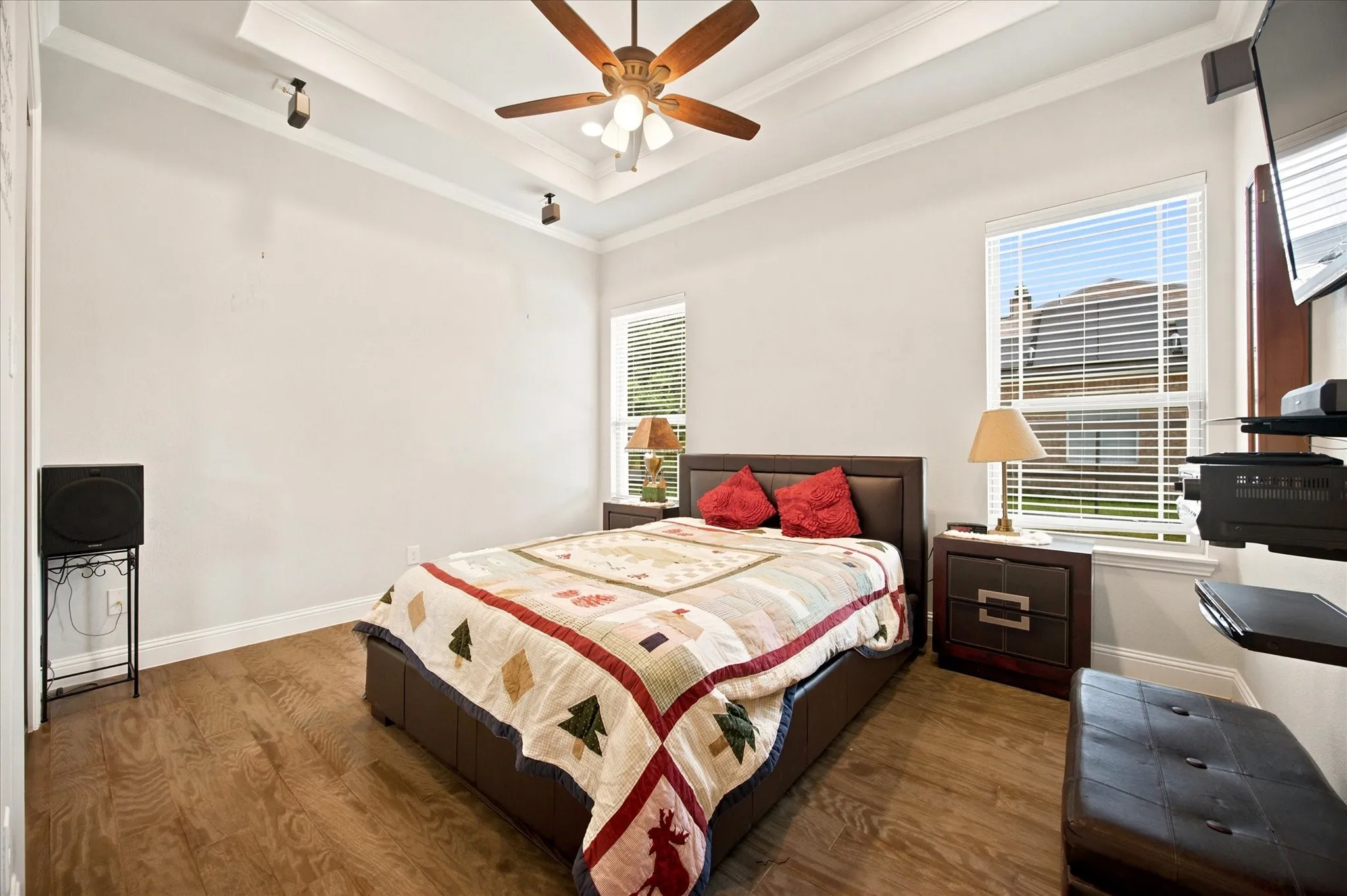Bedroom featuring tray ceiling, crown molding, wood-finish flooring, two windows with blinds, and a ceiling fan with integrated lighting