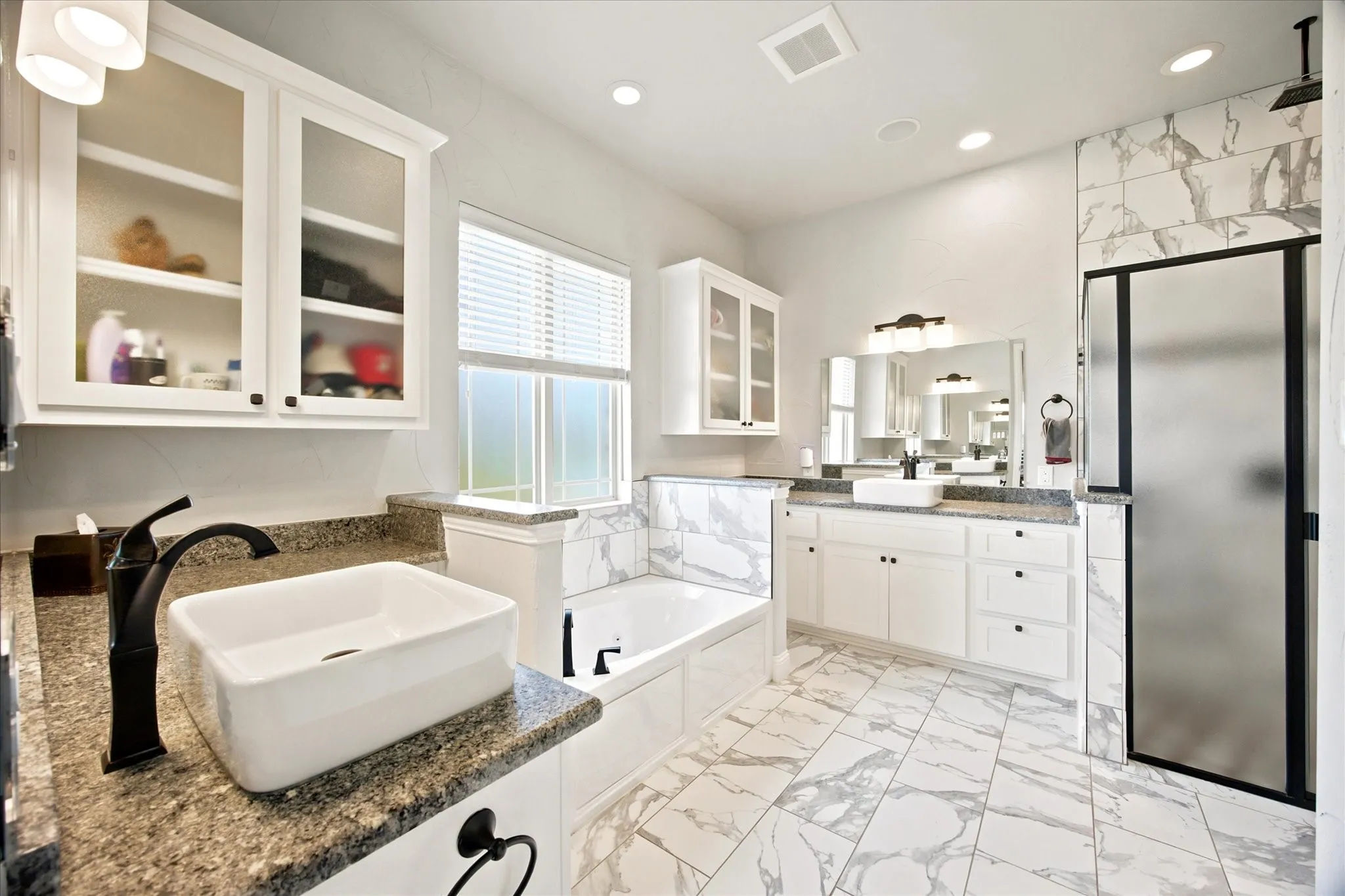 Spacious bathroom featuring a soaking tub, a large walk-in shower with frosted glass, a dual vanity with vessel sinks, white cabinetry, and marble-patterned floor tiling