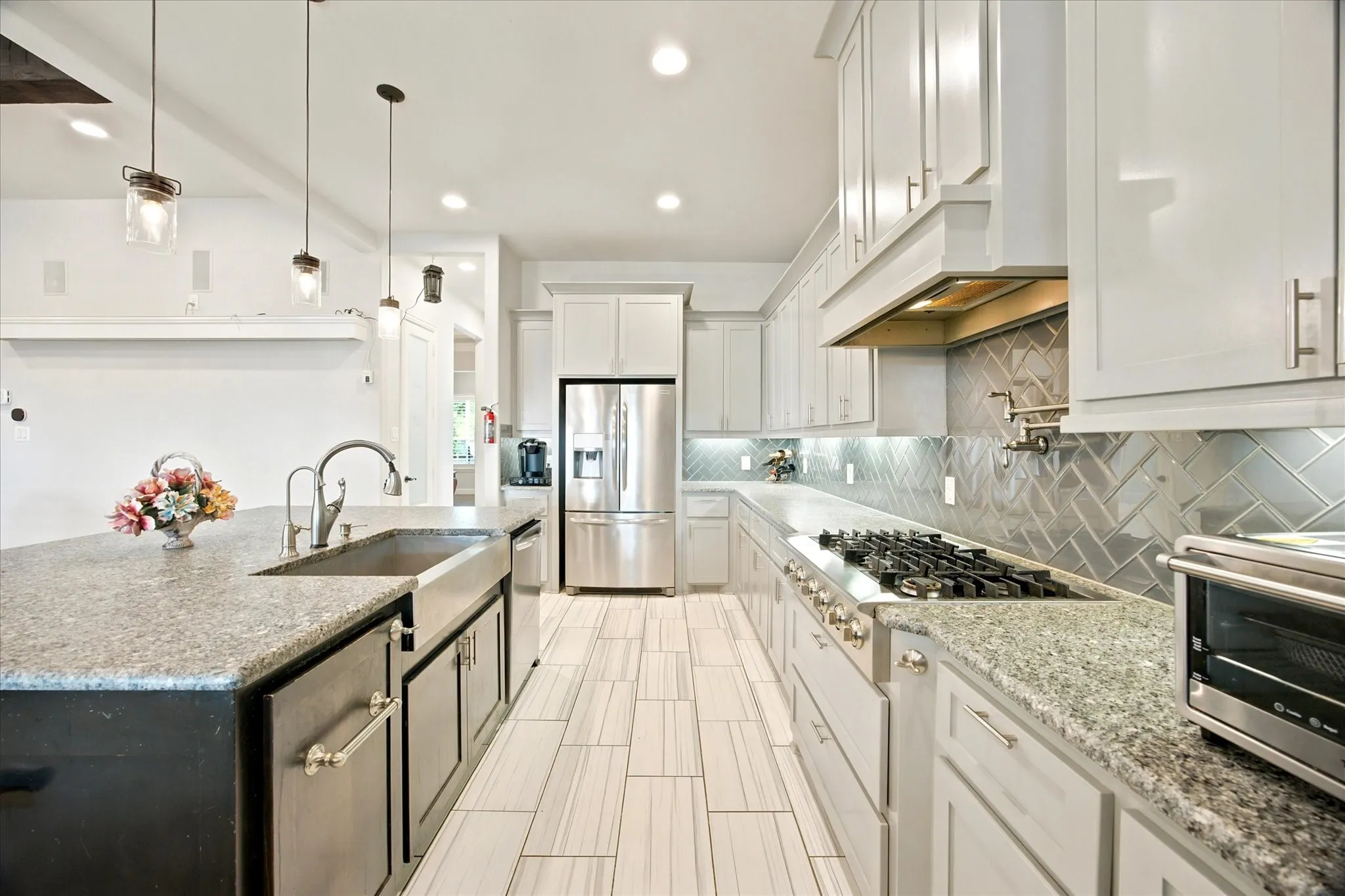 Spacious kitchen featuring an expansive island with a farmhouse sink, light-toned cabinetry, granite countertops, a herringbone tile backsplash, and stainless steel appliances