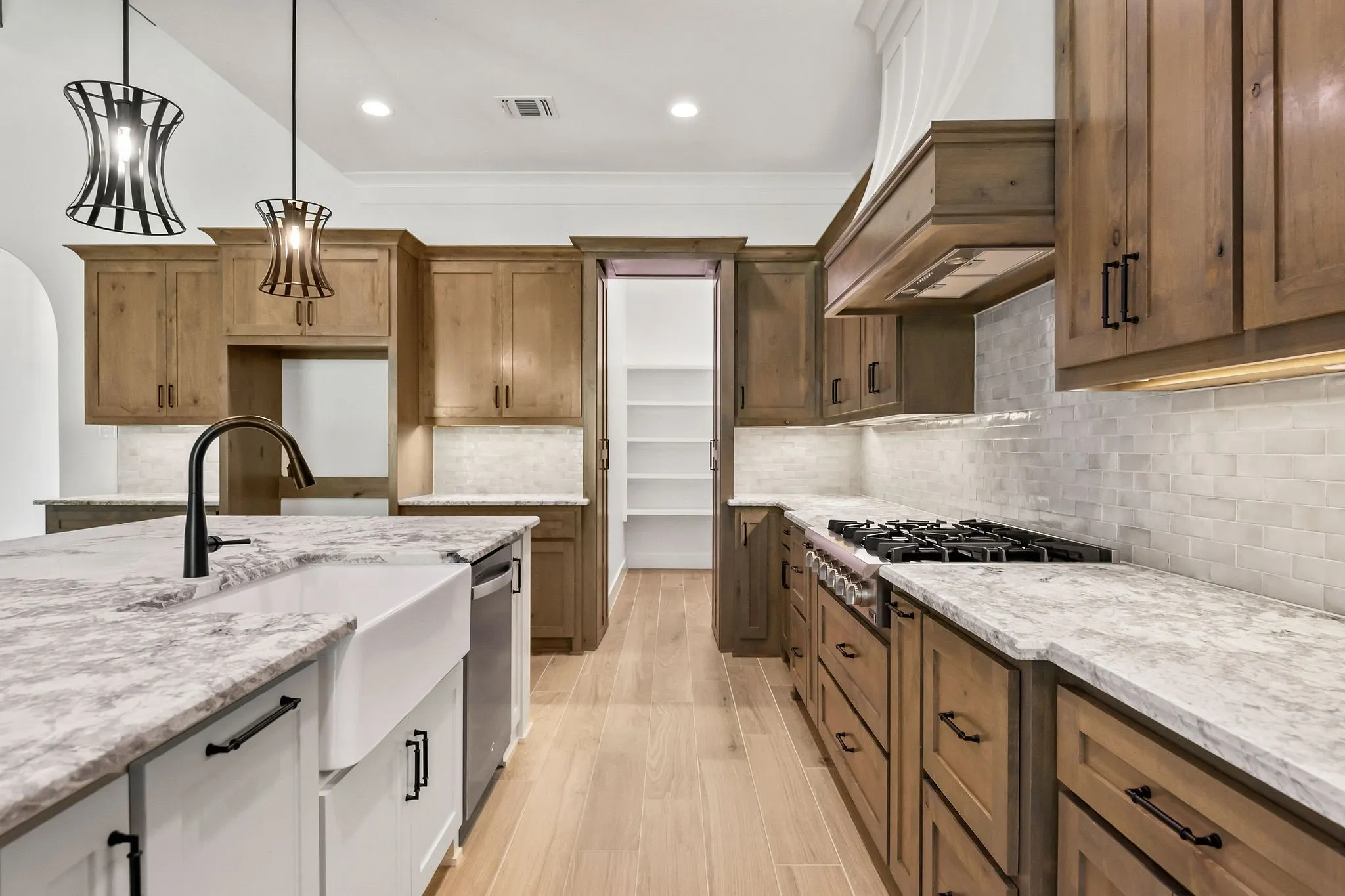 Spacious kitchen featuring an expansive island with a farmhouse sink, stainless steel dishwasher, wood-finish cabinetry, light-toned countertops, and a full tile backsplash