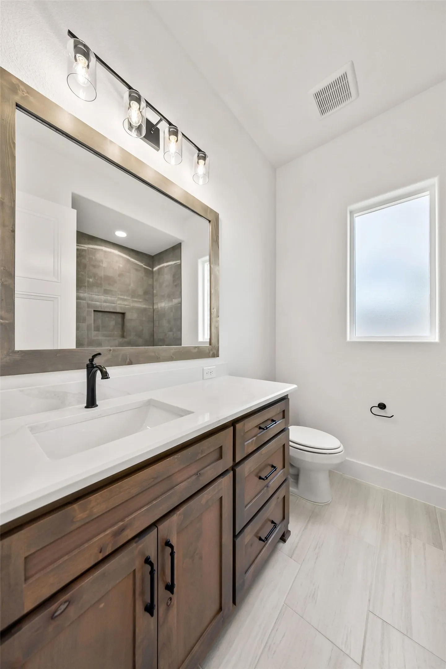 Bathroom featuring a wood-finish vanity with a white countertop, an integrated rectangular sink, a black wall-mounted faucet, and a framed mirror with a four-bulb vanity light fixture