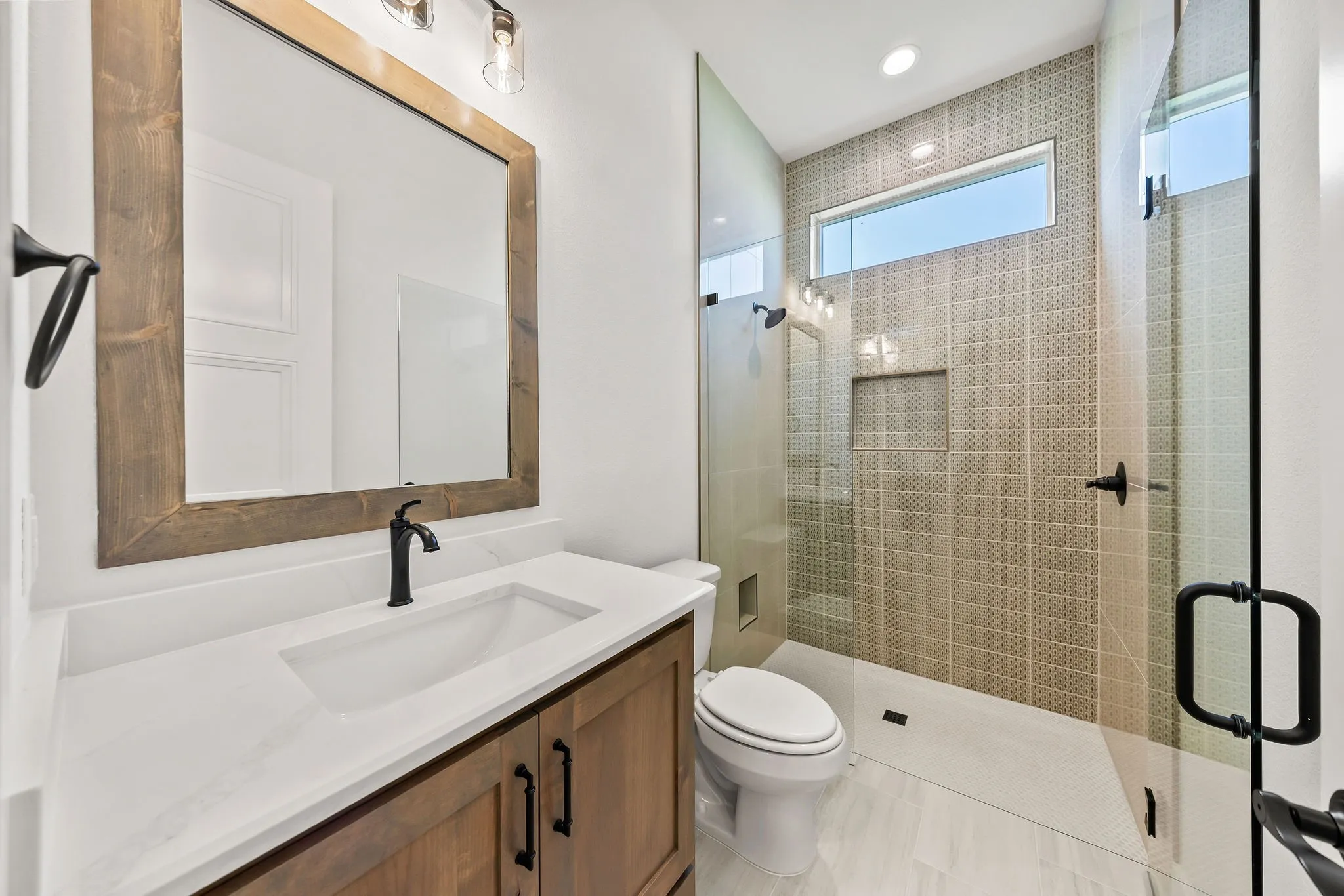 Contemporary bathroom featuring a wood-finish vanity with a white countertop and integrated sink, a frameless wood-frame mirror, and a walk-in shower with mosaic tile walls and a horizontal privacy window
