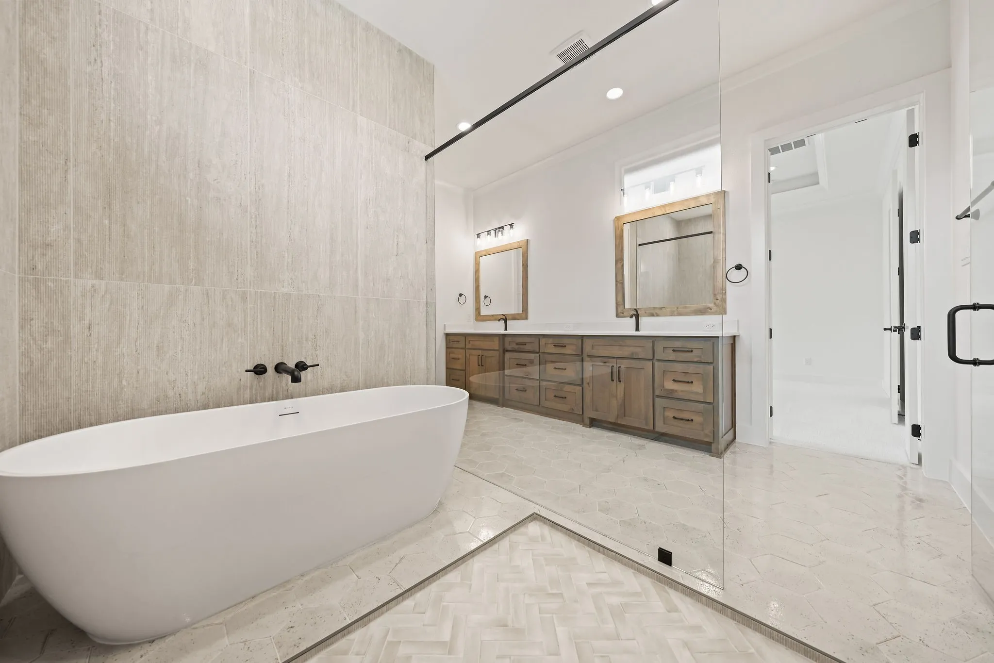 Contemporary bathroom featuring a freestanding soaking tub, dual wood-finish vanity with stone countertops, oversized wall-mounted mirrors, and a spacious glass-enclosed shower with herringbone tile flooring