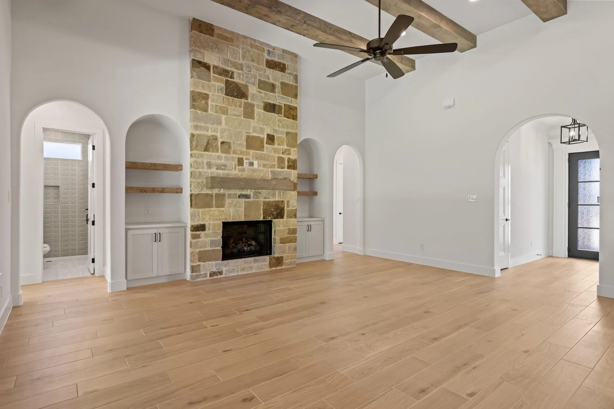 Spacious room featuring light wood-finish flooring, a stone fireplace with a substantial hearth, built-in cabinetry with floating shelves, and exposed ceiling beams
