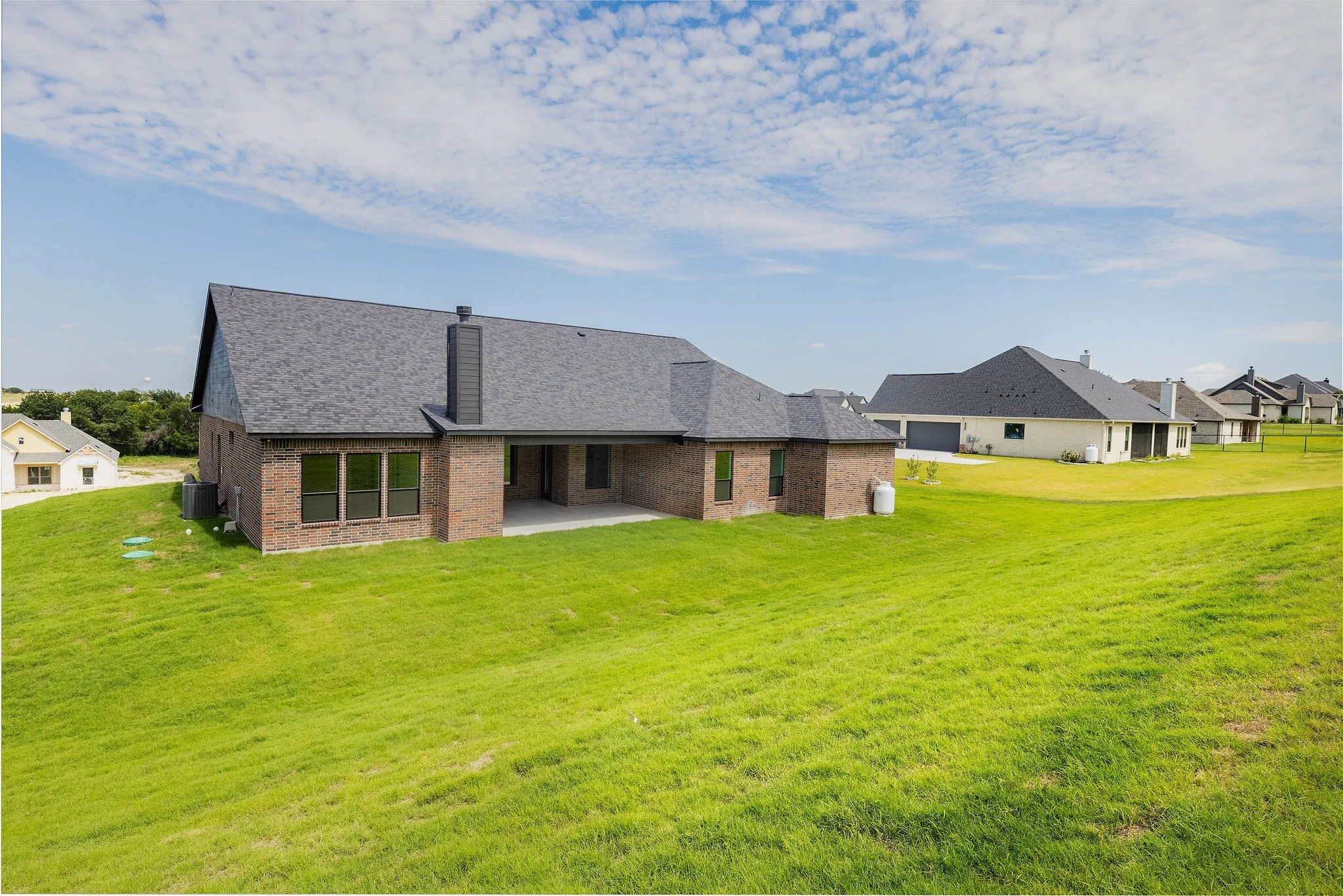Brick exterior with a dark shingle roof, featuring multiple windows, a covered patio, and a sloped grass yard