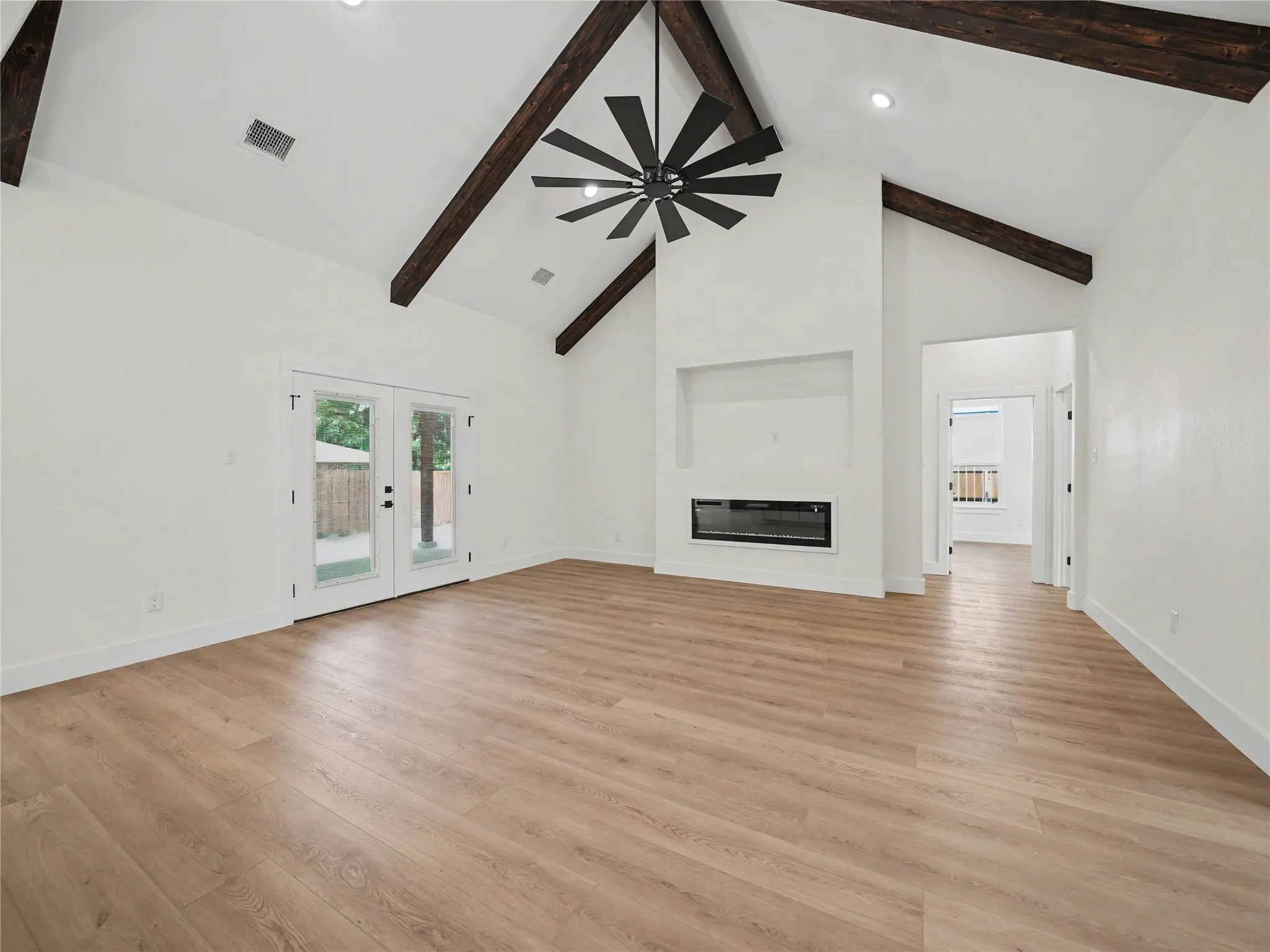 Vaulted ceilings with exposed dark wood beams, contemporary ceiling fan, recessed lighting, integrated linear fireplace, and wood-finish flooring