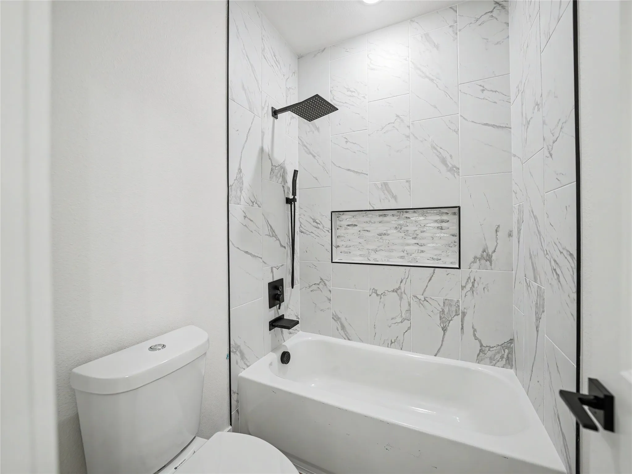 Modern bathroom featuring a bathtub with a rainfall showerhead, white marble-look tile surround, recessed tile niche, and matte black fixtures