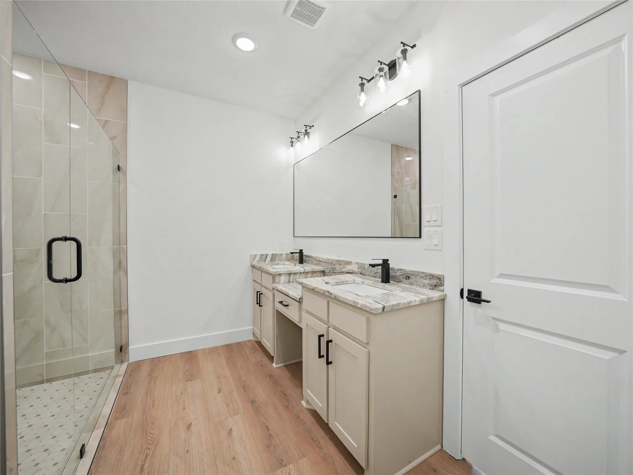 Bathroom featuring a frameless glass shower enclosure with tiled walls and a mosaic floor, a dual vanity with light-toned cabinetry, granite countertops, and matte black fixtures, wood-finish flooring, and recessed lighting