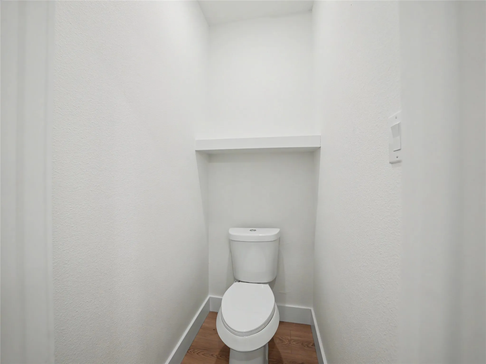 Recessed toilet alcove featuring a built-in white shelf, white baseboards, and wood-finish flooring