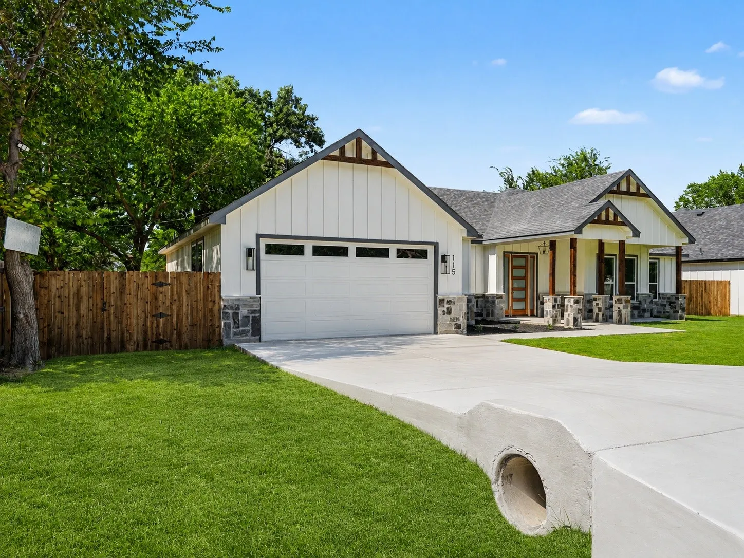 Modern ranch-style home featuring white board and batten siding, dark stone accents, wood-look porch columns, a gray shingled roof, and a concrete driveway