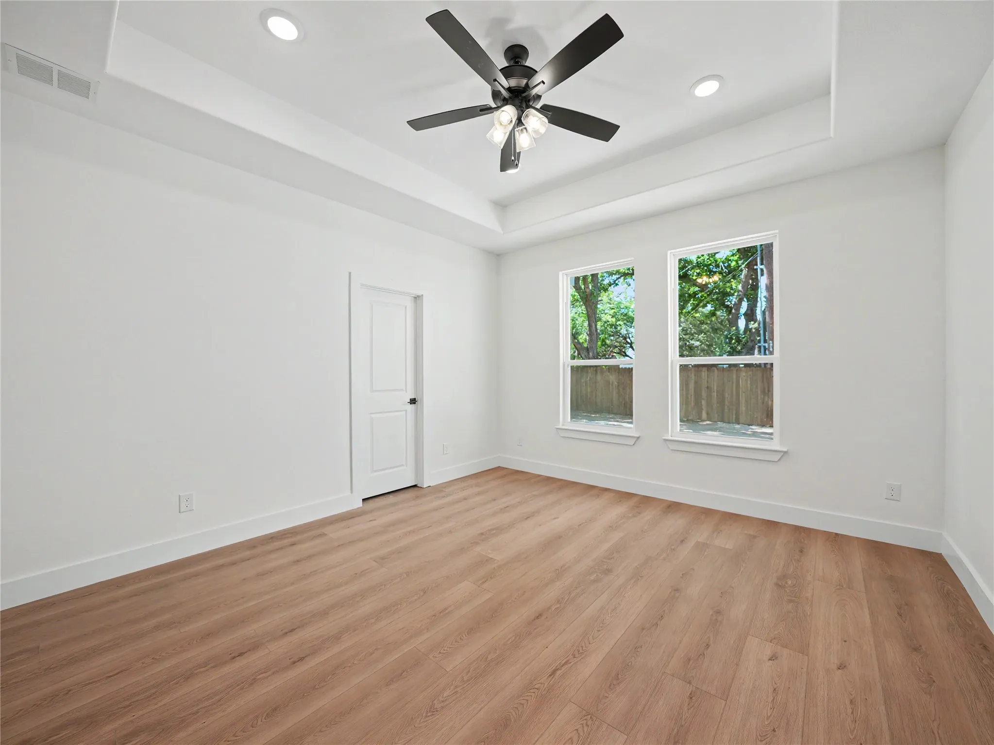 Spacious room featuring wood-finish flooring, two windows, a tray ceiling with recessed lighting, and a ceiling fan with integrated lighting