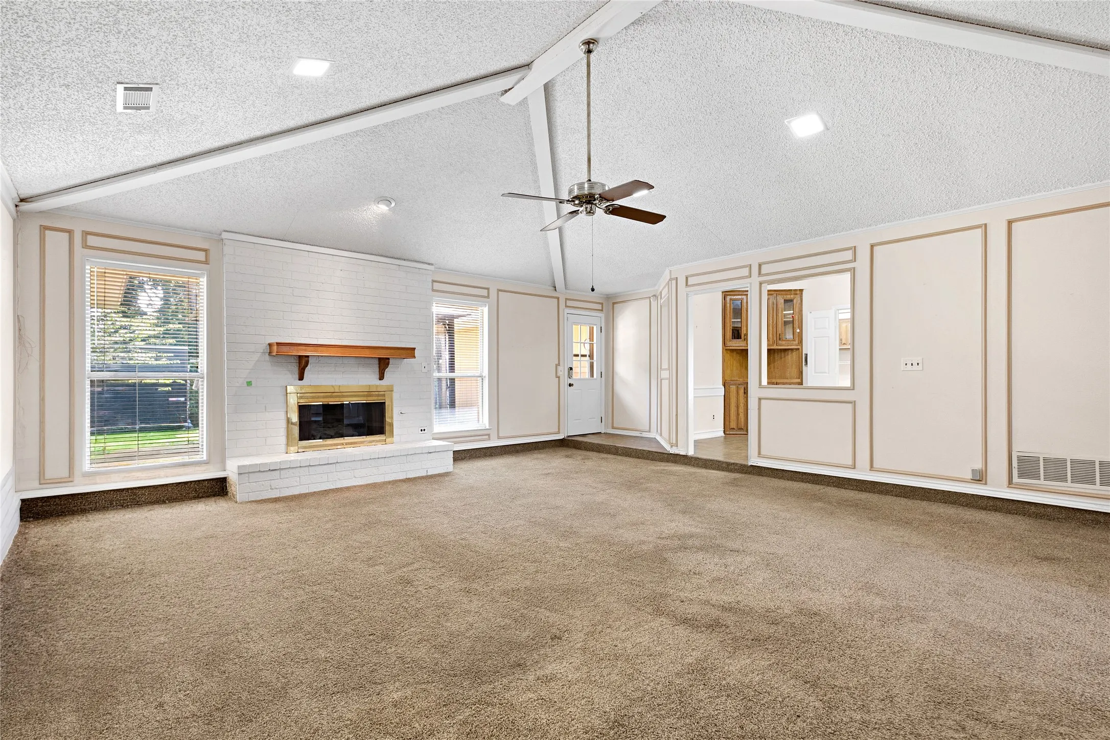 Spacious living area featuring a vaulted ceiling with exposed beams, an exposed brick fireplace with a wood mantle, and a ceiling fan