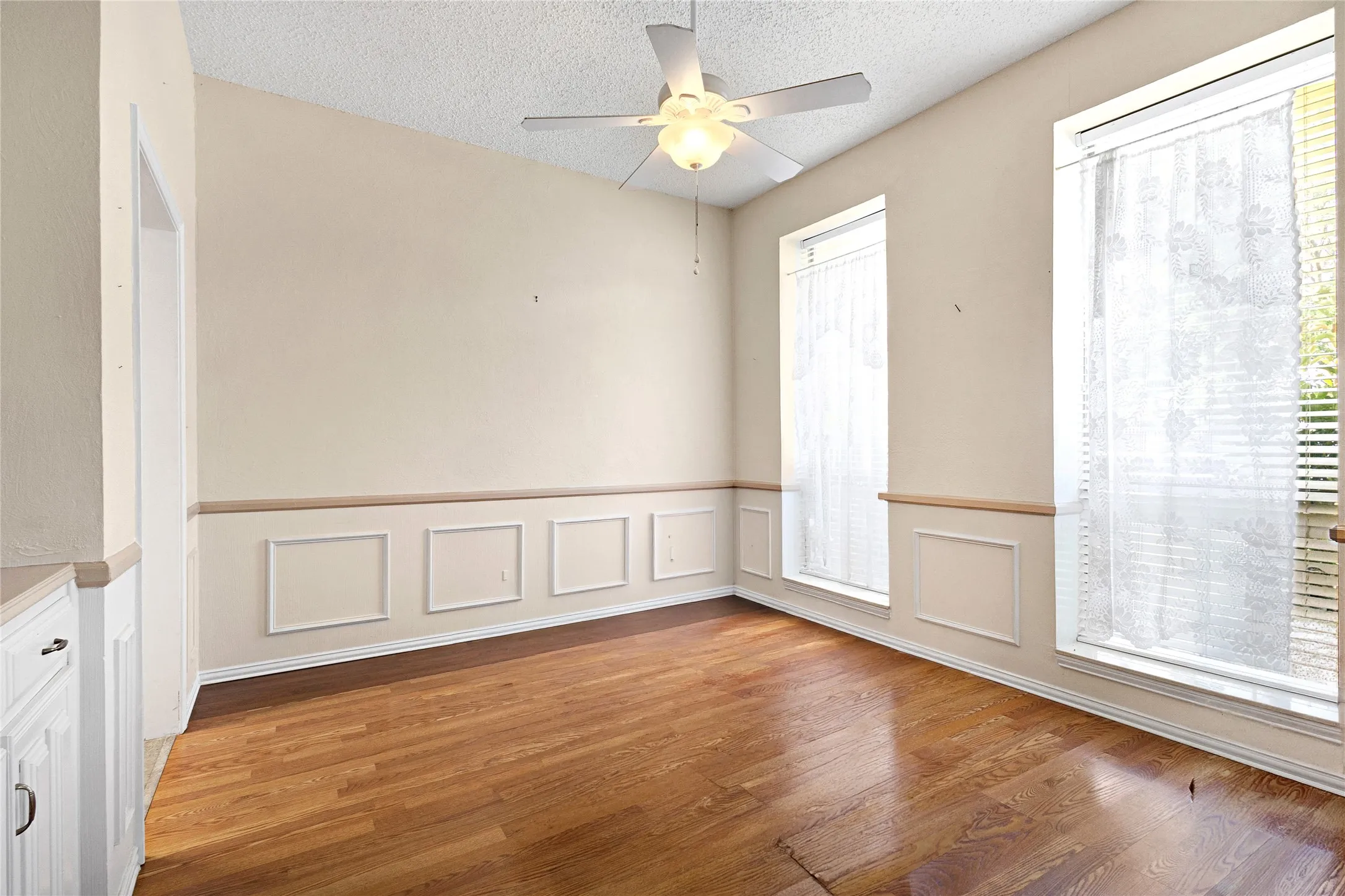 Spacious room featuring wood-finish flooring, two large windows with blinds, a ceiling fan, and decorative wainscoting