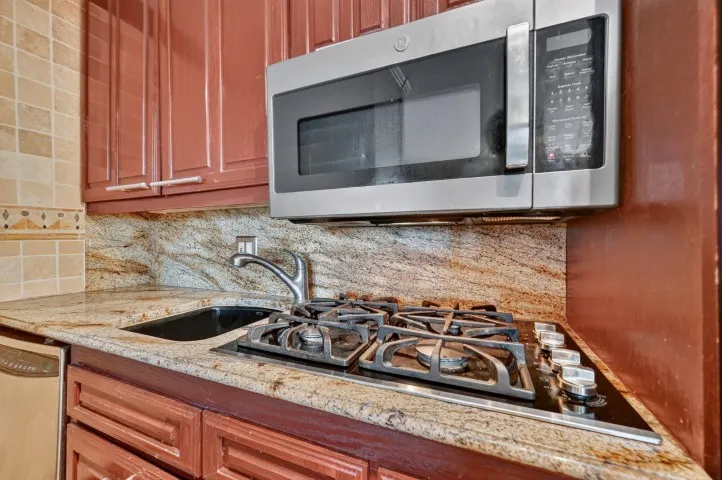 Kitchen featuring granite countertops, wood-finish cabinetry, stainless steel microwave, gas cooktop, and an undermount sink with a gooseneck faucet