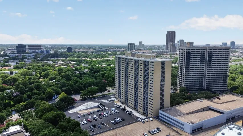 Aerial perspective showcasing two high-rise buildings with a visible parking area, surrounded by a verdant urban landscape