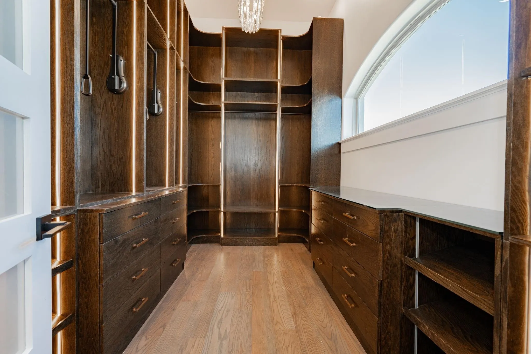 Custom walk-in closet featuring dark wood cabinetry, integrated lighting, extensive shelving, drawer storage, and a large arched window