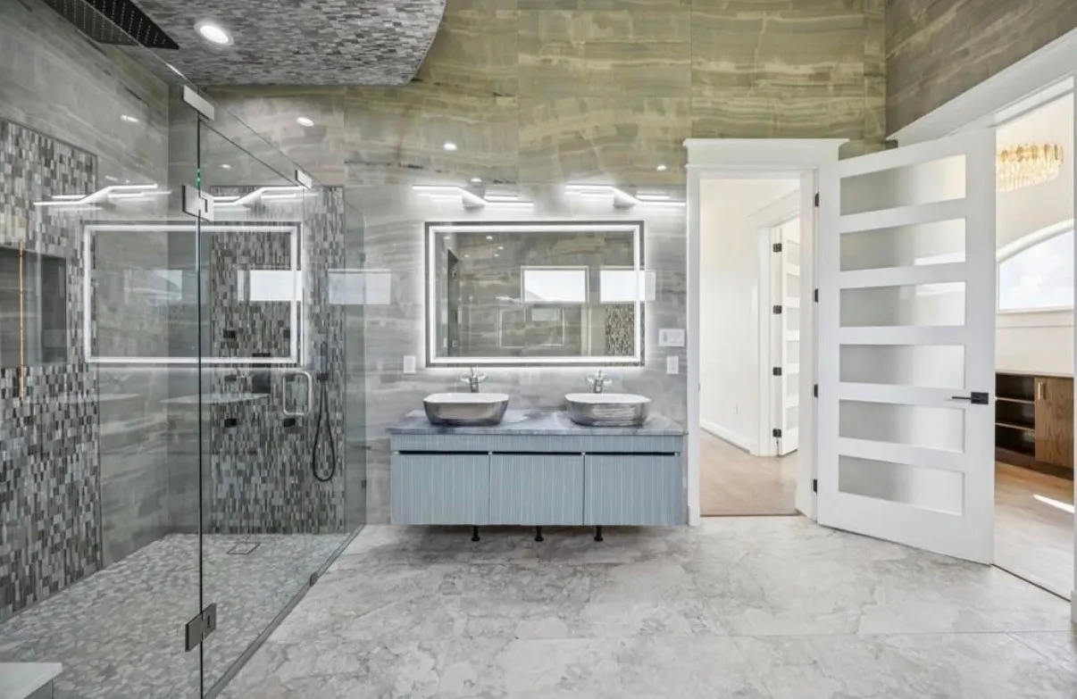 Spacious bathroom featuring a glass-enclosed shower with mosaic tile accents, a floating double vanity with vessel sinks, back-lit mirrors, and large format floor tiling
