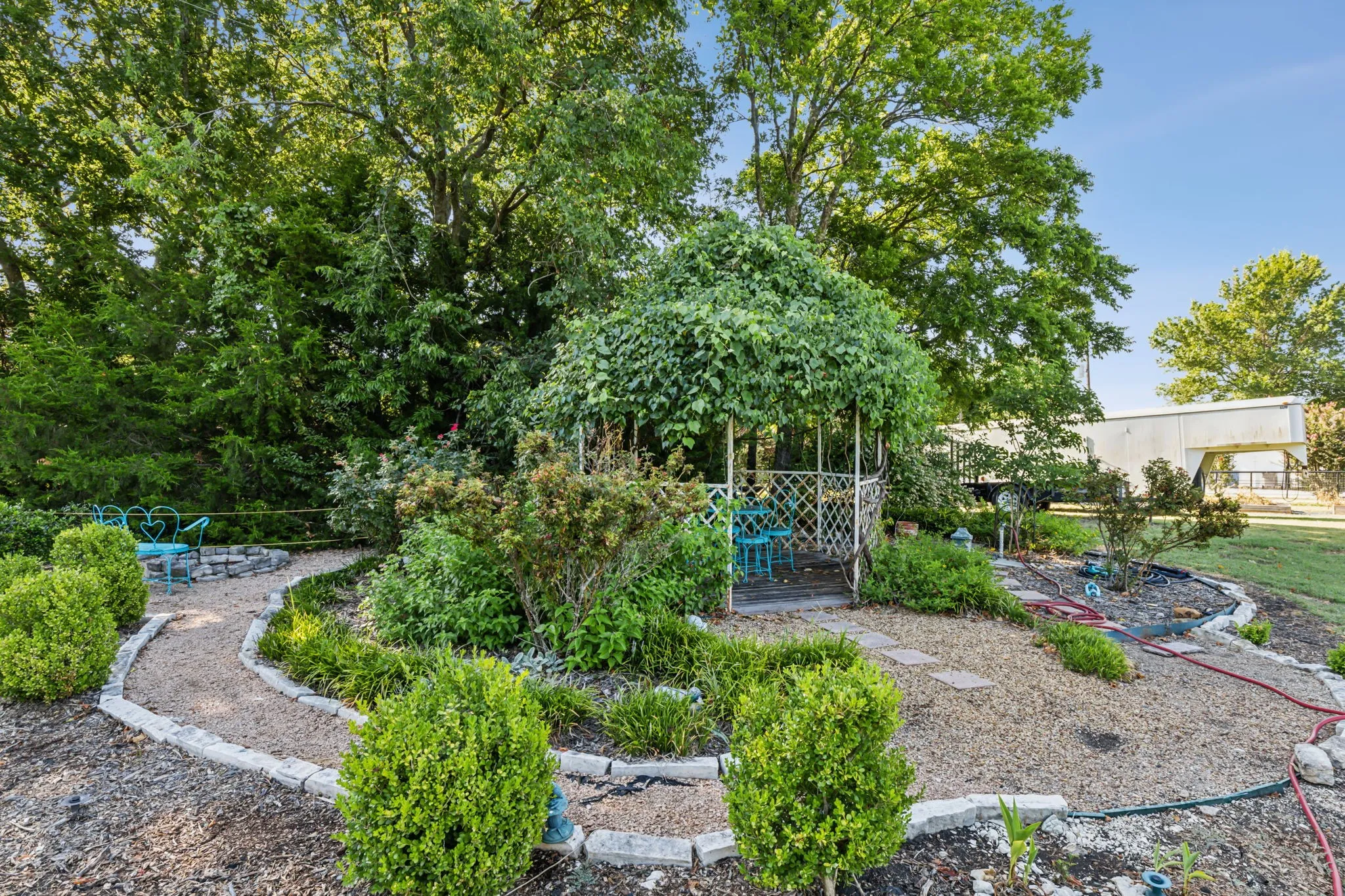 Landscaped garden featuring a central gazebo with climbing foliage, a gravel pathway with stone edging, and various shrubs and trees