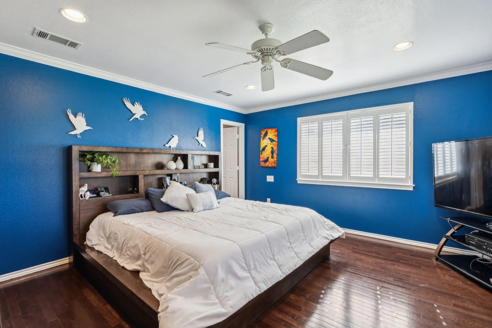 Bedroom featuring dark wood-finish flooring, white crown molding, a ceiling fan with integrated lighting, recessed ceiling lights, and a window with white plantation shutters