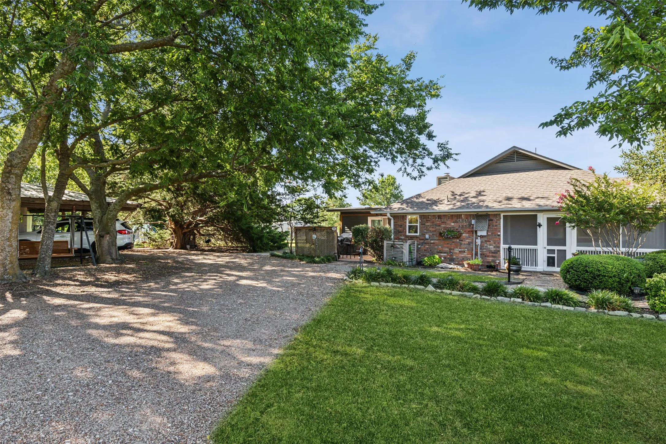 Rear exterior featuring a brick facade, screened porch, mature landscaping, and a gravel driveway leading to a covered parking area
