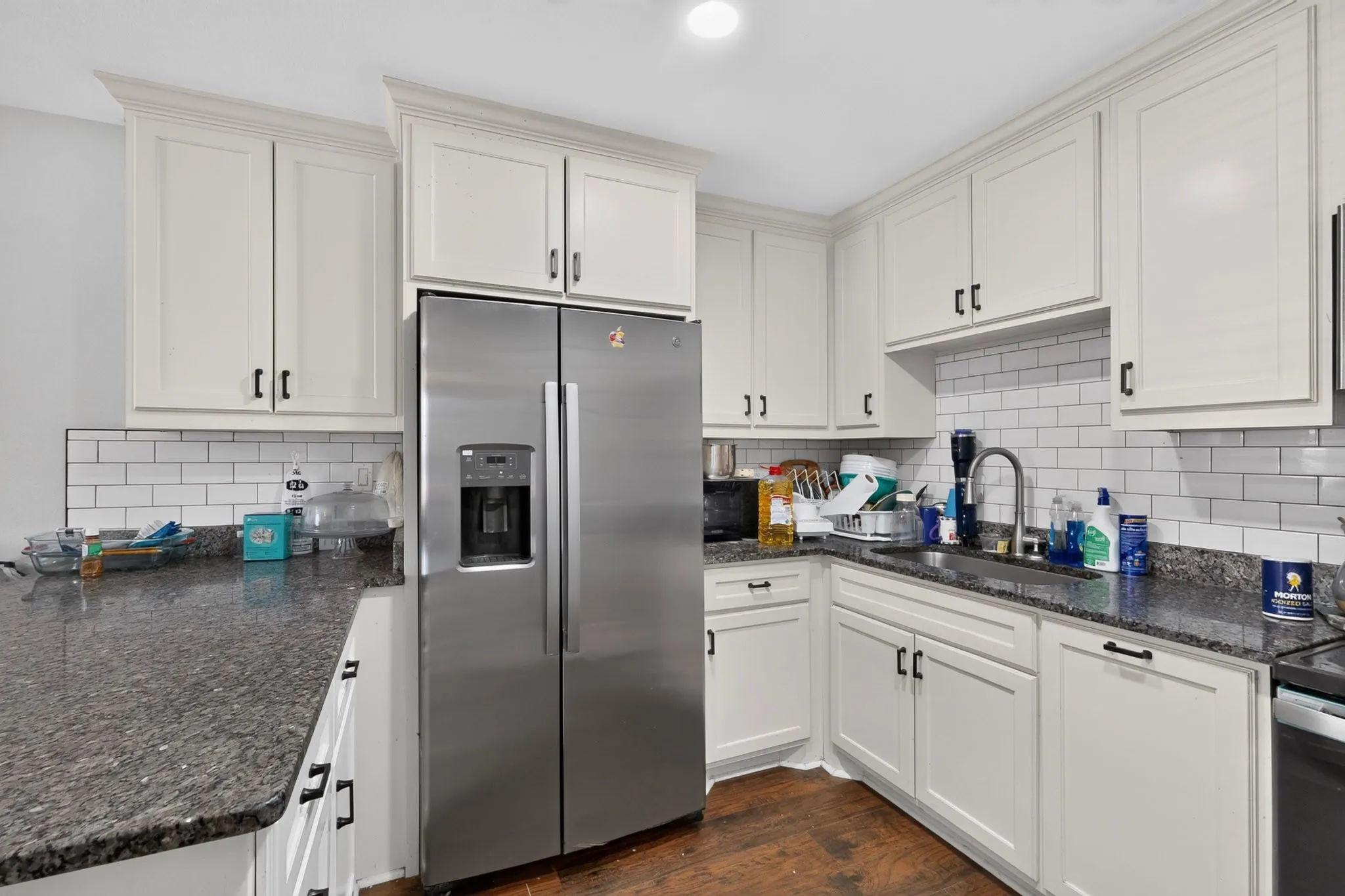 Kitchen featuring white shaker-style cabinetry, dark speckled countertops, subway tile backsplash, stainless steel side-by-side refrigerator, and a stainless steel sink with a high-arc faucet